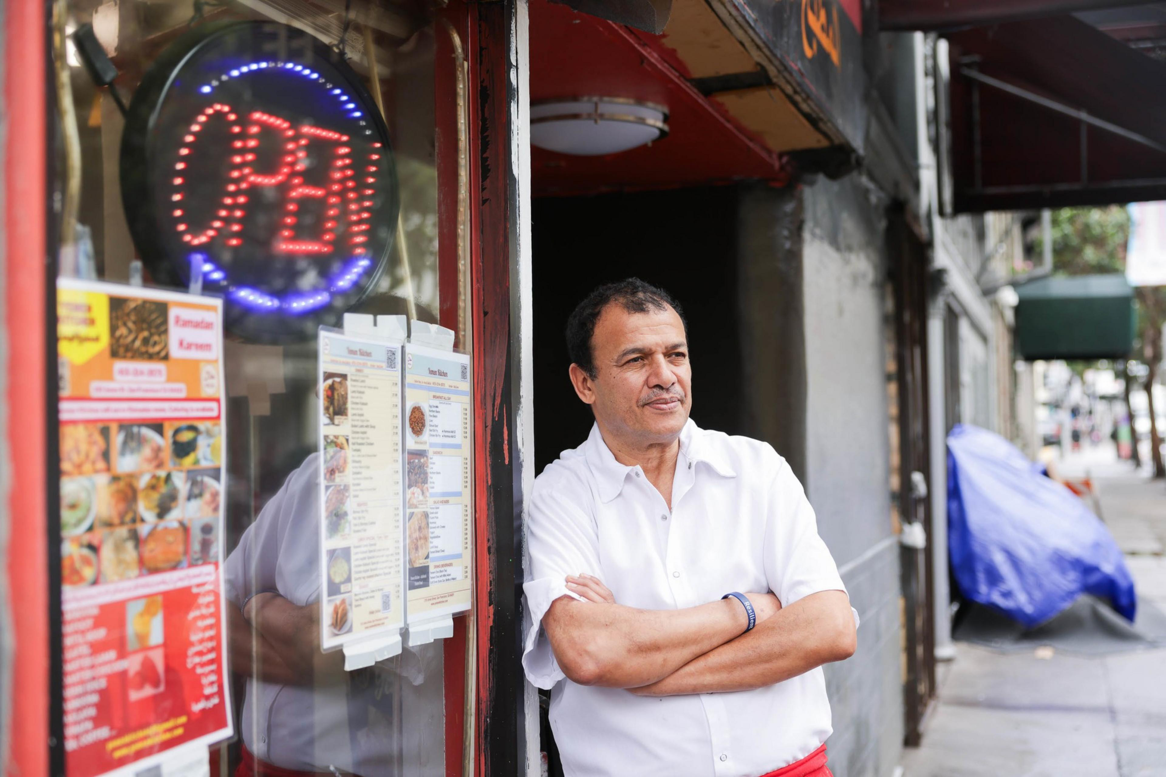 A man stands leaning against a restaurant doorway beneath a neon &quot;OPEN&quot; sign, with menus displayed next to him.