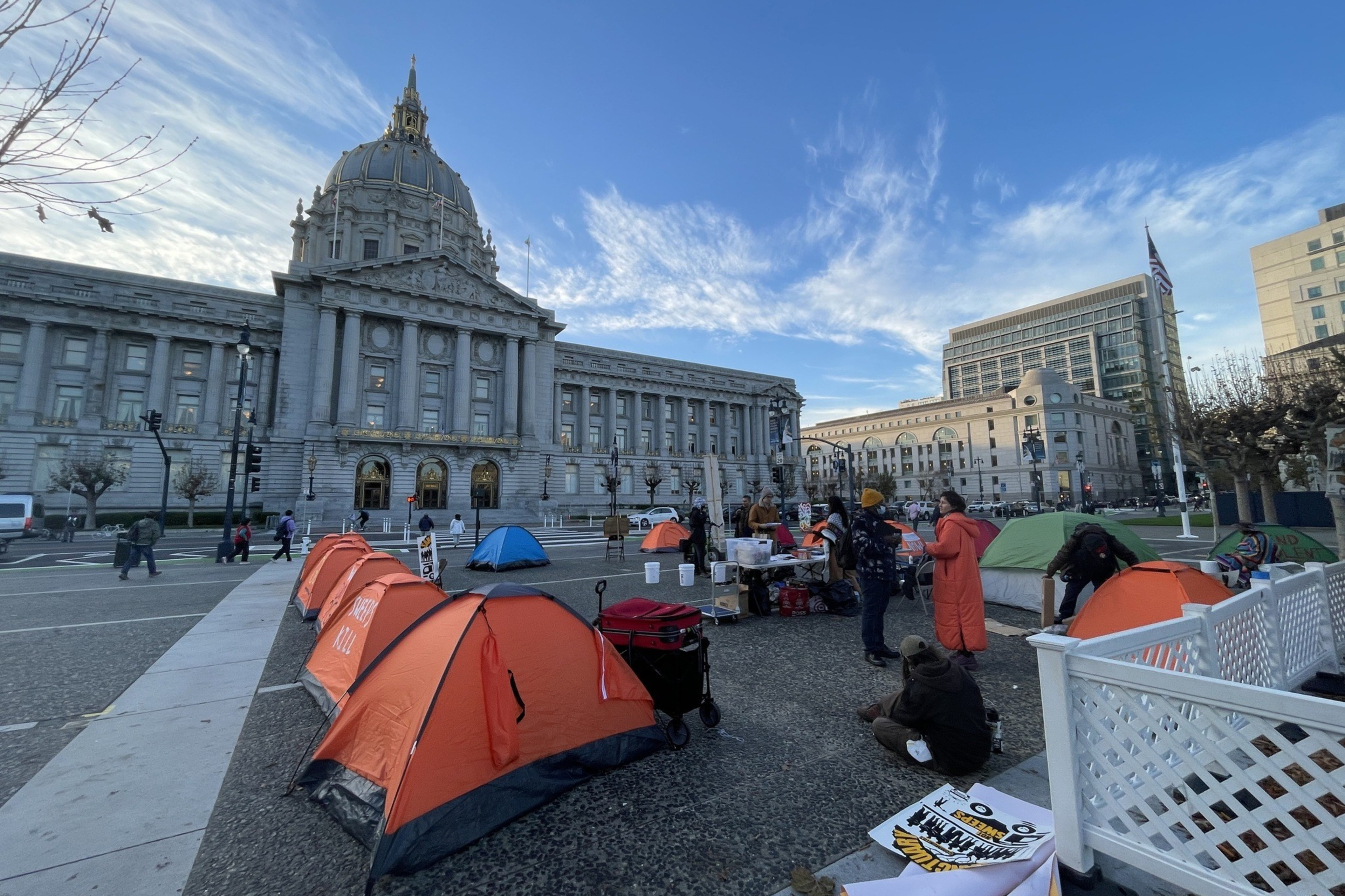People gather around tents set up in front of a large, domed government building. The scene is lively, with a clear sky and modern buildings nearby.