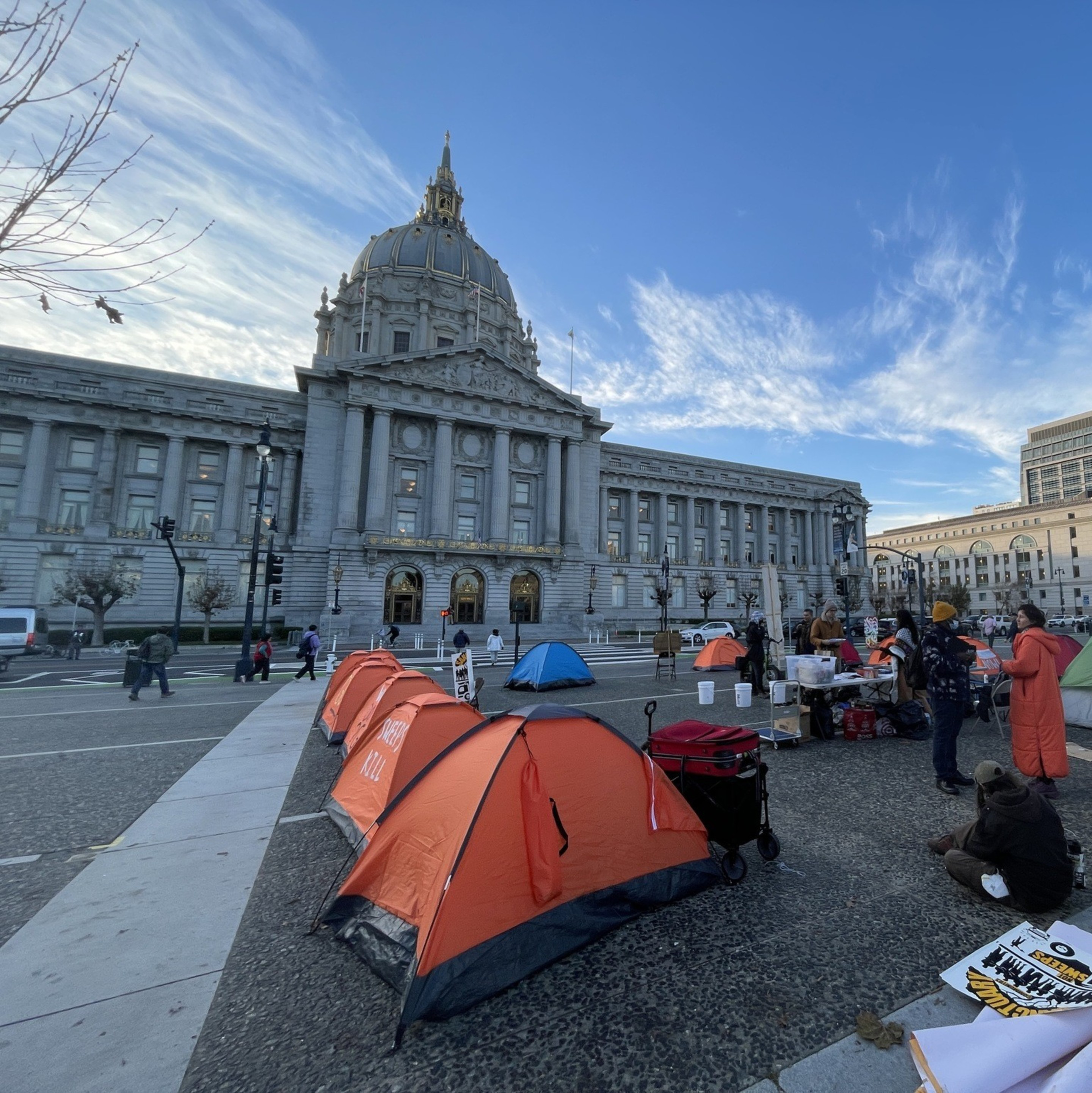 People gather around tents set up in front of a large, domed government building. The scene is lively, with a clear sky and modern buildings nearby.