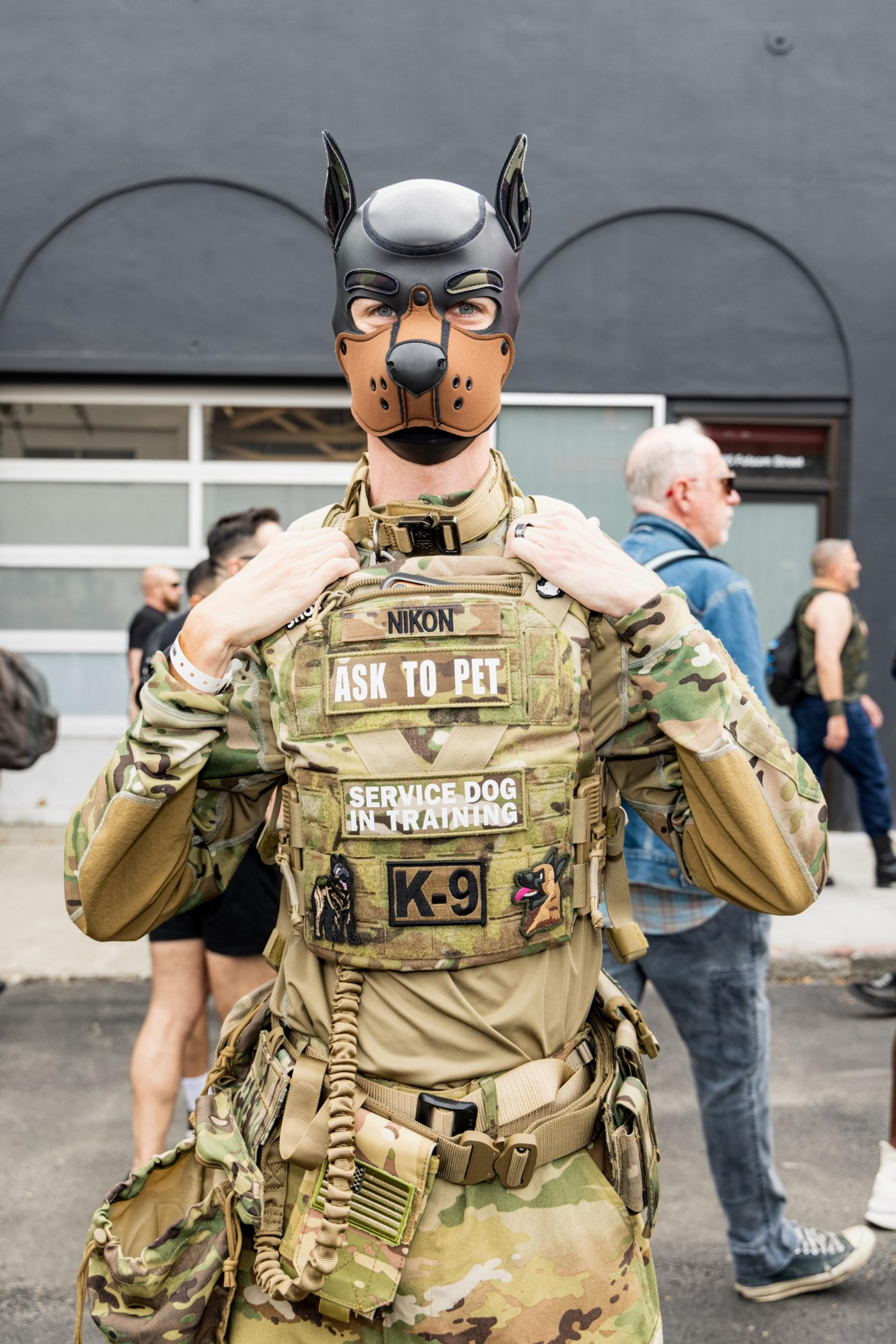 A person in camouflage gear wears a dog mask and a vest labeled “ASK TO PET,” “SERVICE DOG IN TRAINING,” and “K-9,” standing outdoors with people nearby.
