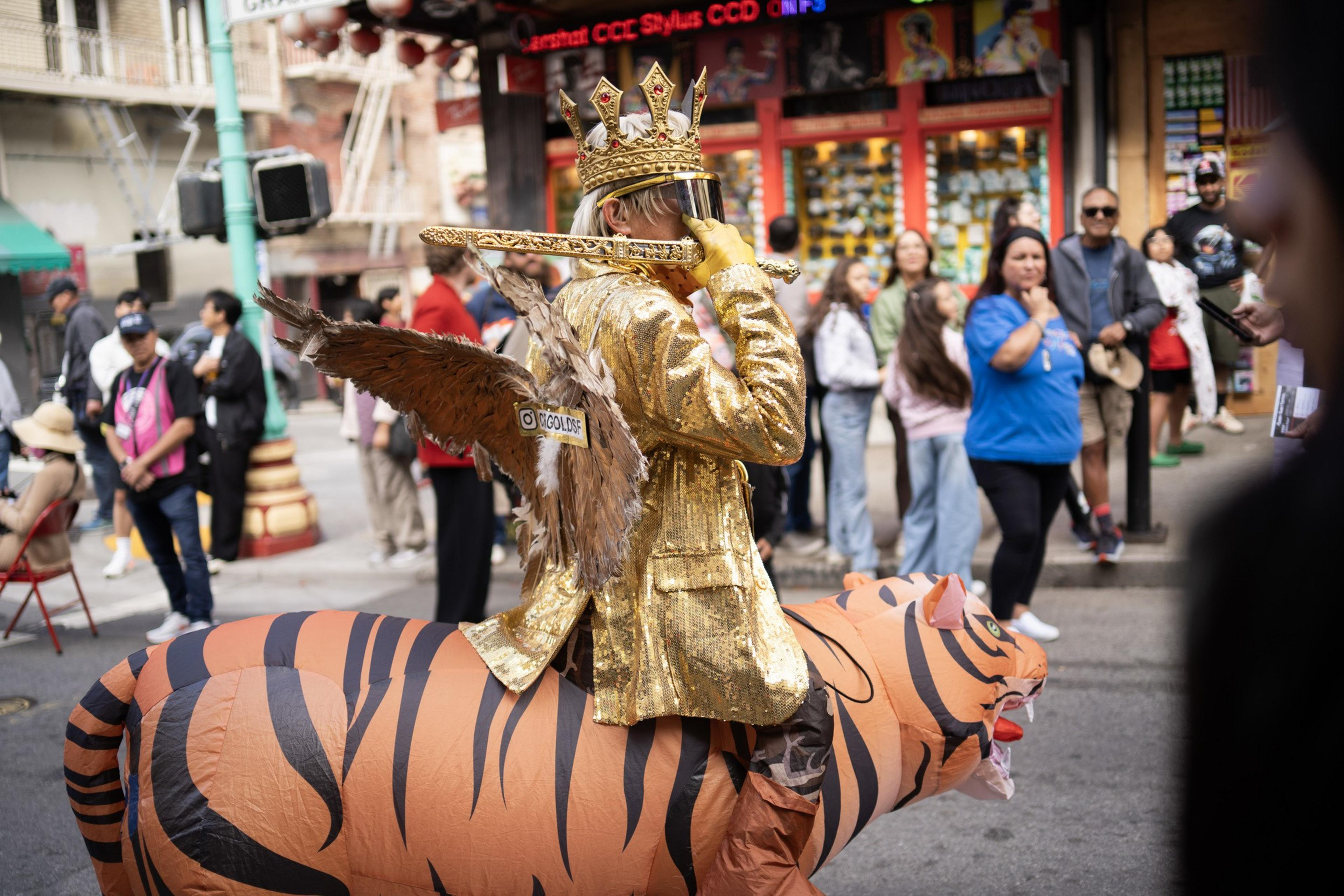 A person wearing a gold crown, gold jacket, and feathered wings plays a flute while riding an inflatable tiger costume on a busy street.