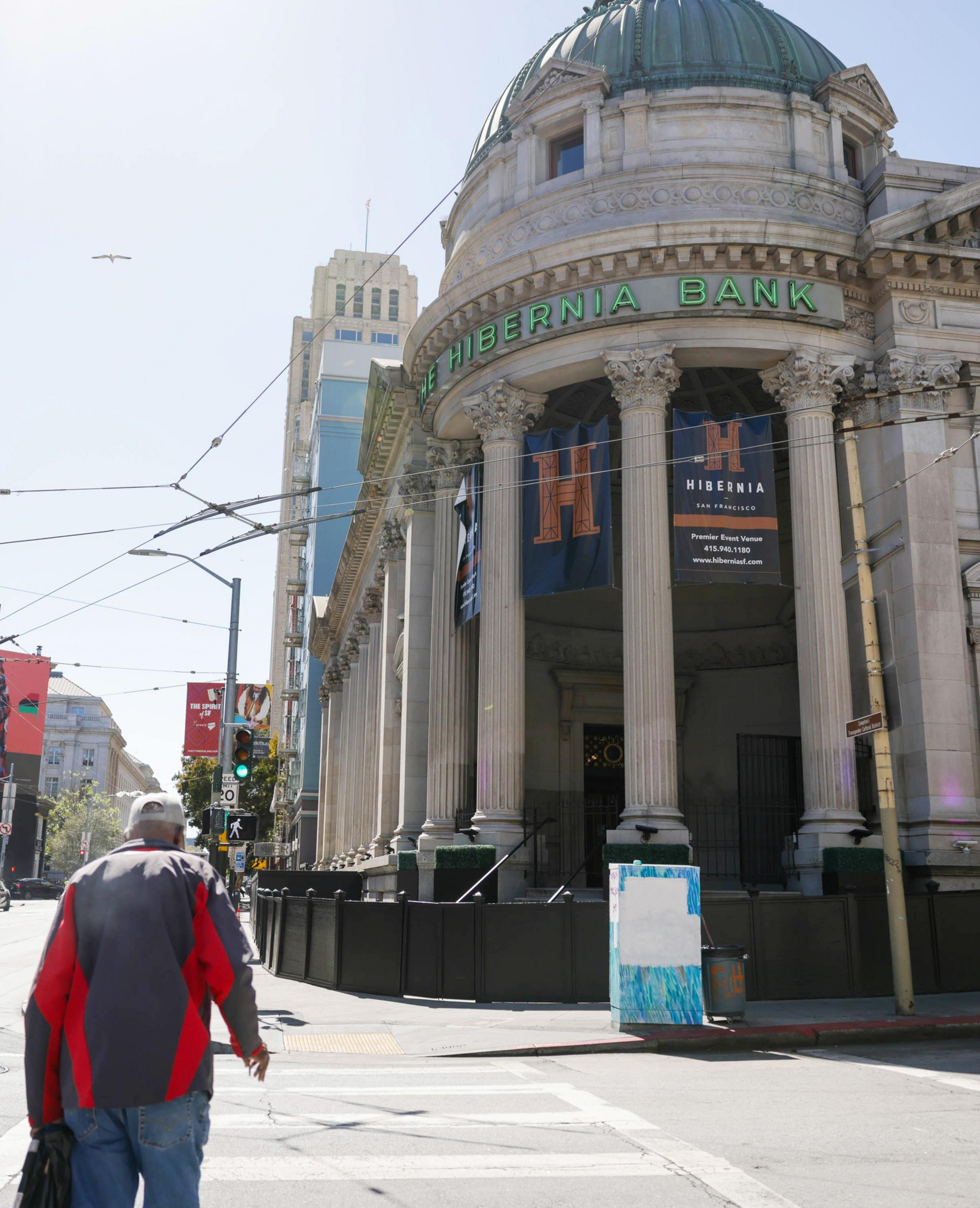 A person walks by the Hibernia Bank with a domed roof and classical architecture on a sunny day with a clear sky.
