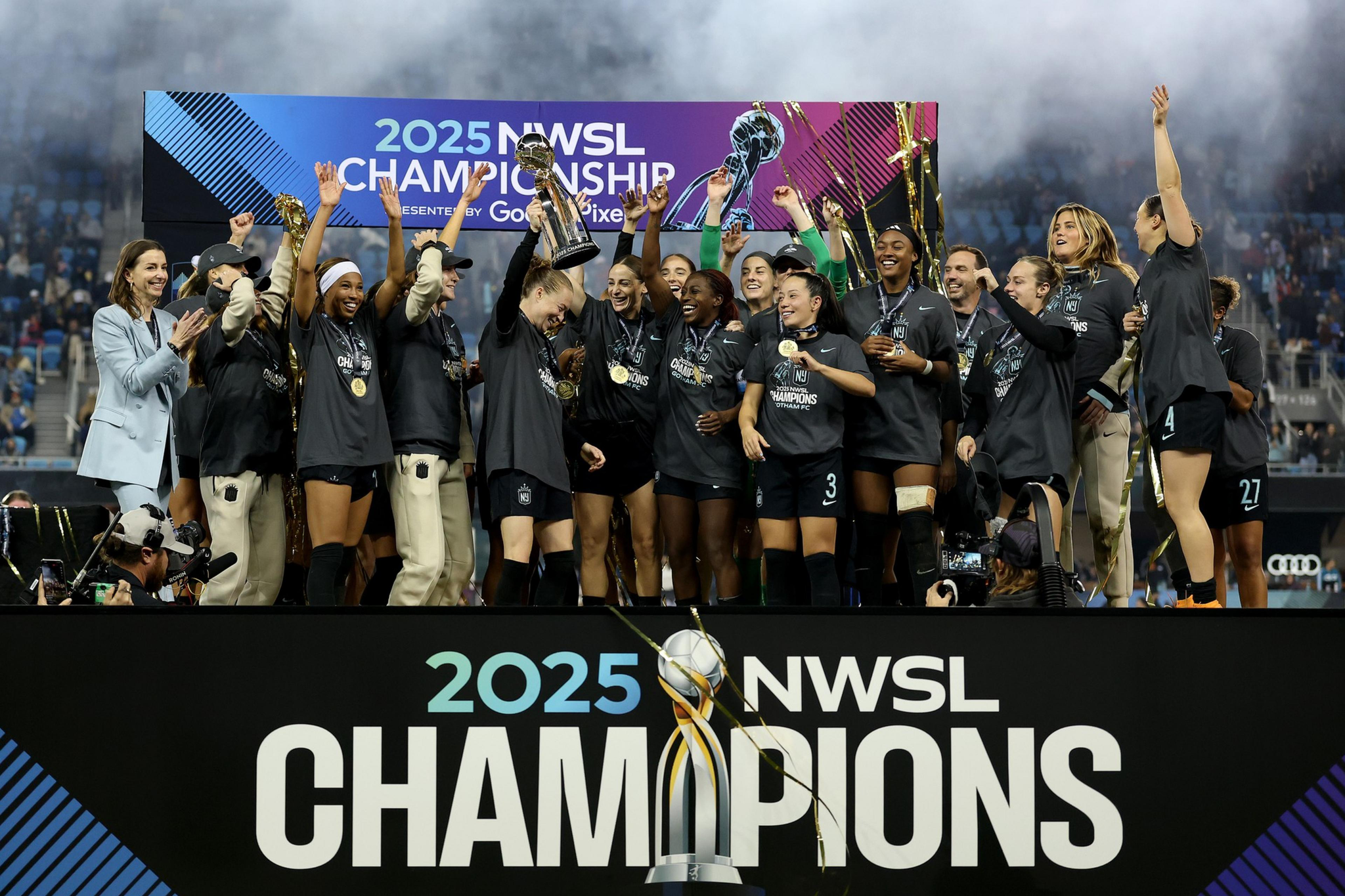 A women's soccer team celebrates on a stage holding a trophy with "2025 NWSL Champions" displayed, wearing medals and cheering joyfully.