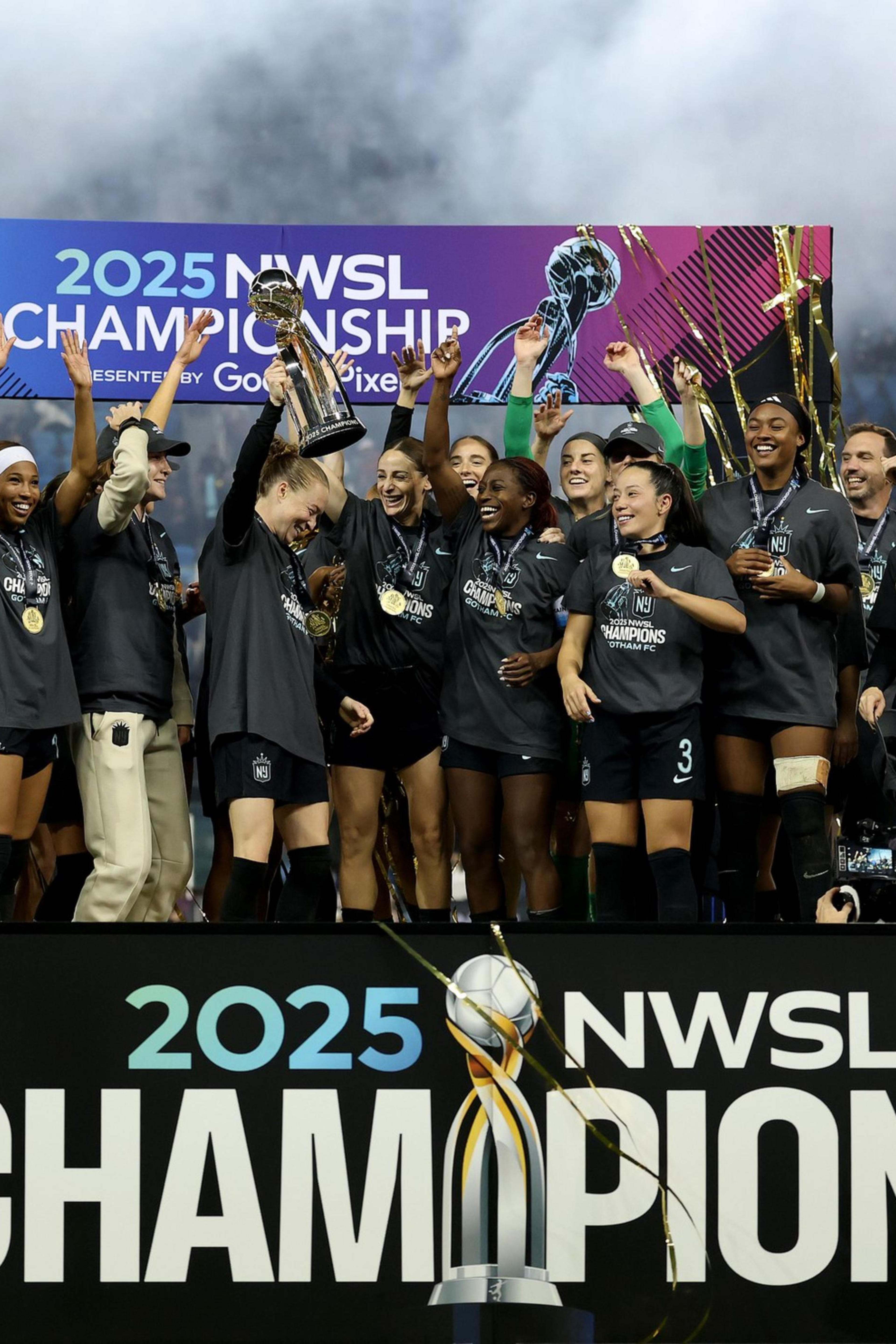A women's soccer team celebrates on a stage holding a trophy with "2025 NWSL Champions" displayed, wearing medals and cheering joyfully.