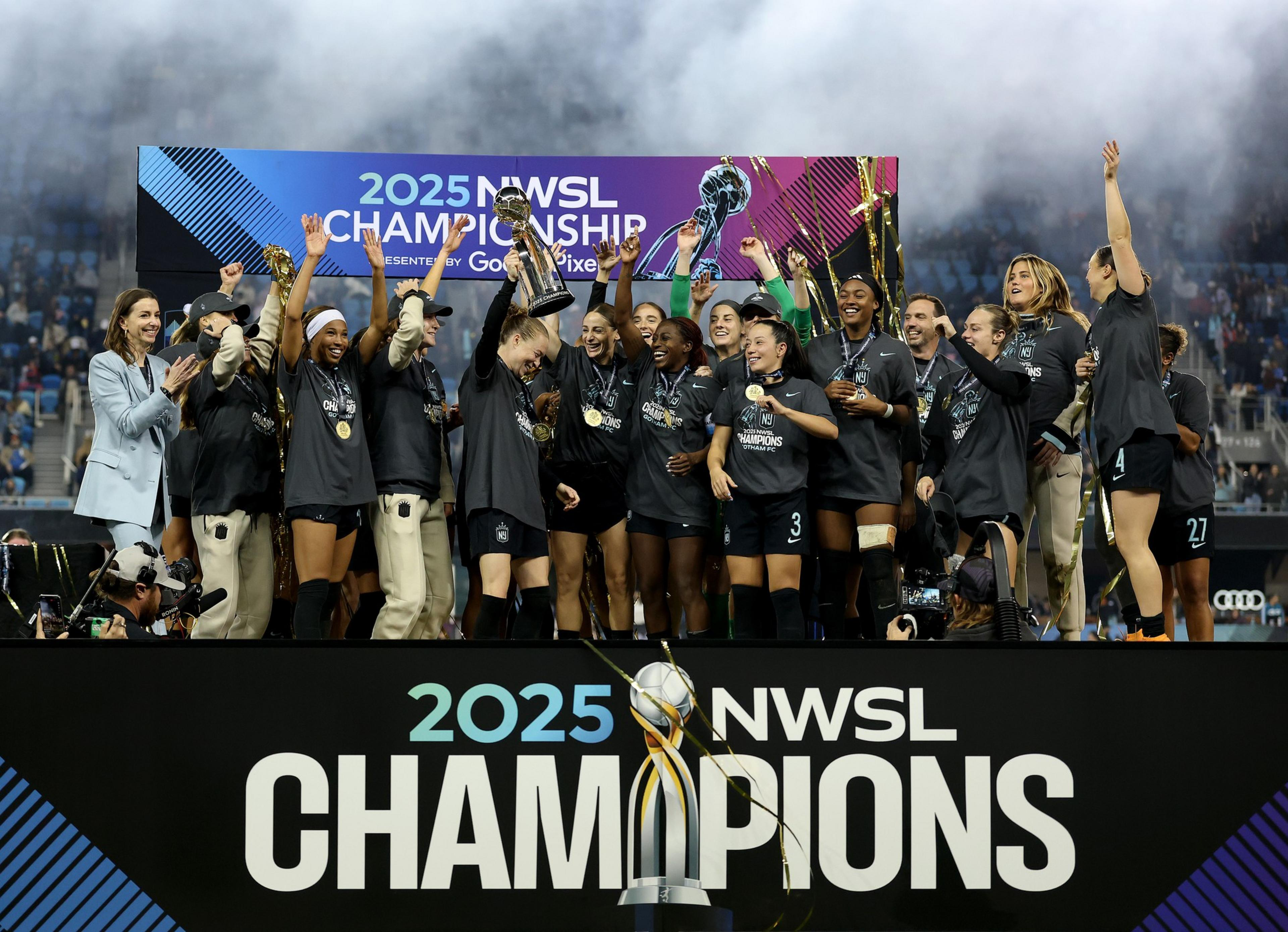 A women's soccer team celebrates on a stage holding a trophy with "2025 NWSL Champions" displayed, wearing medals and cheering joyfully.
