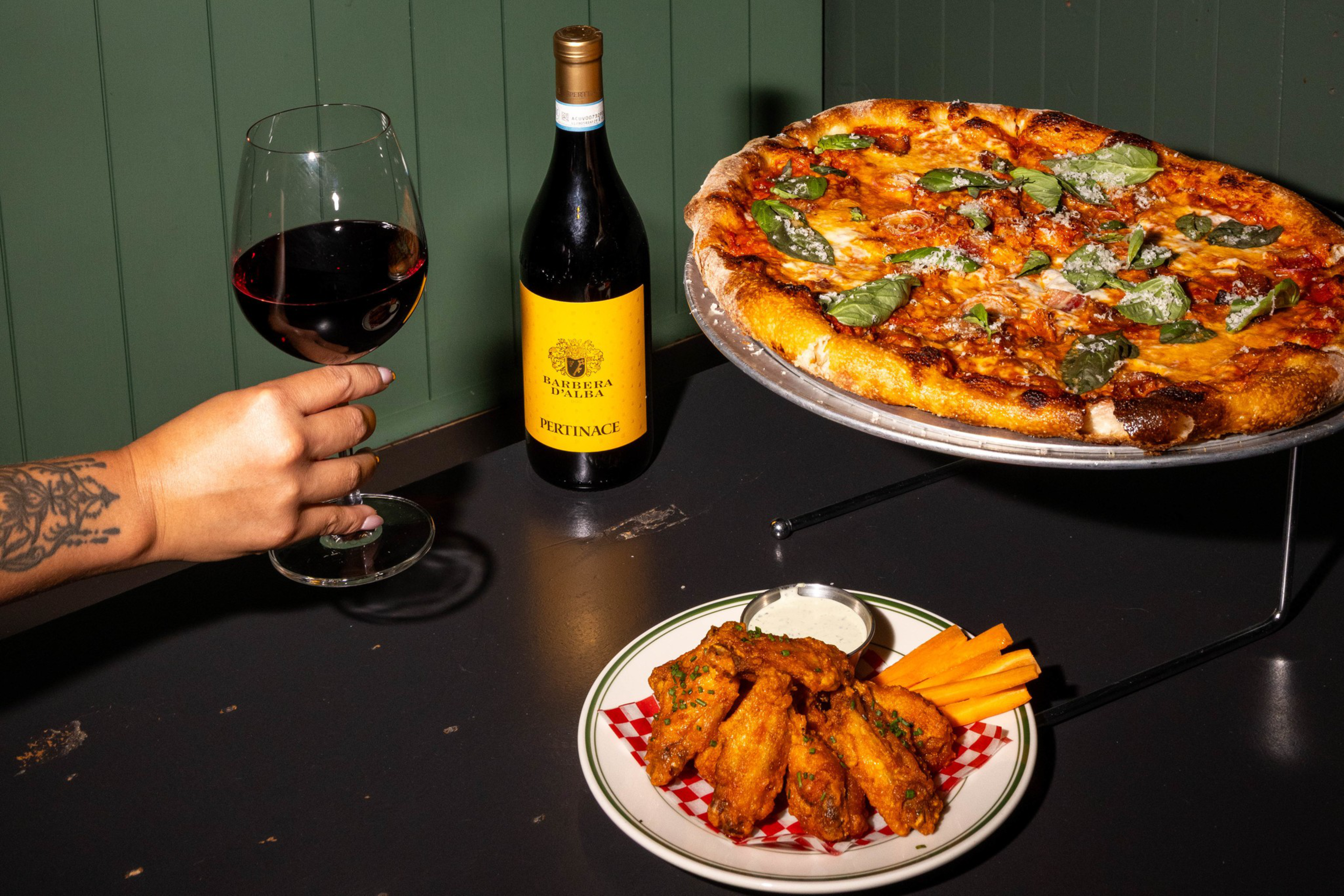 A hand holding a glass of red wine near a bottle of wine, a plate of chicken wings with carrot sticks and dip, and a pizza topped with fresh basil on a raised stand.