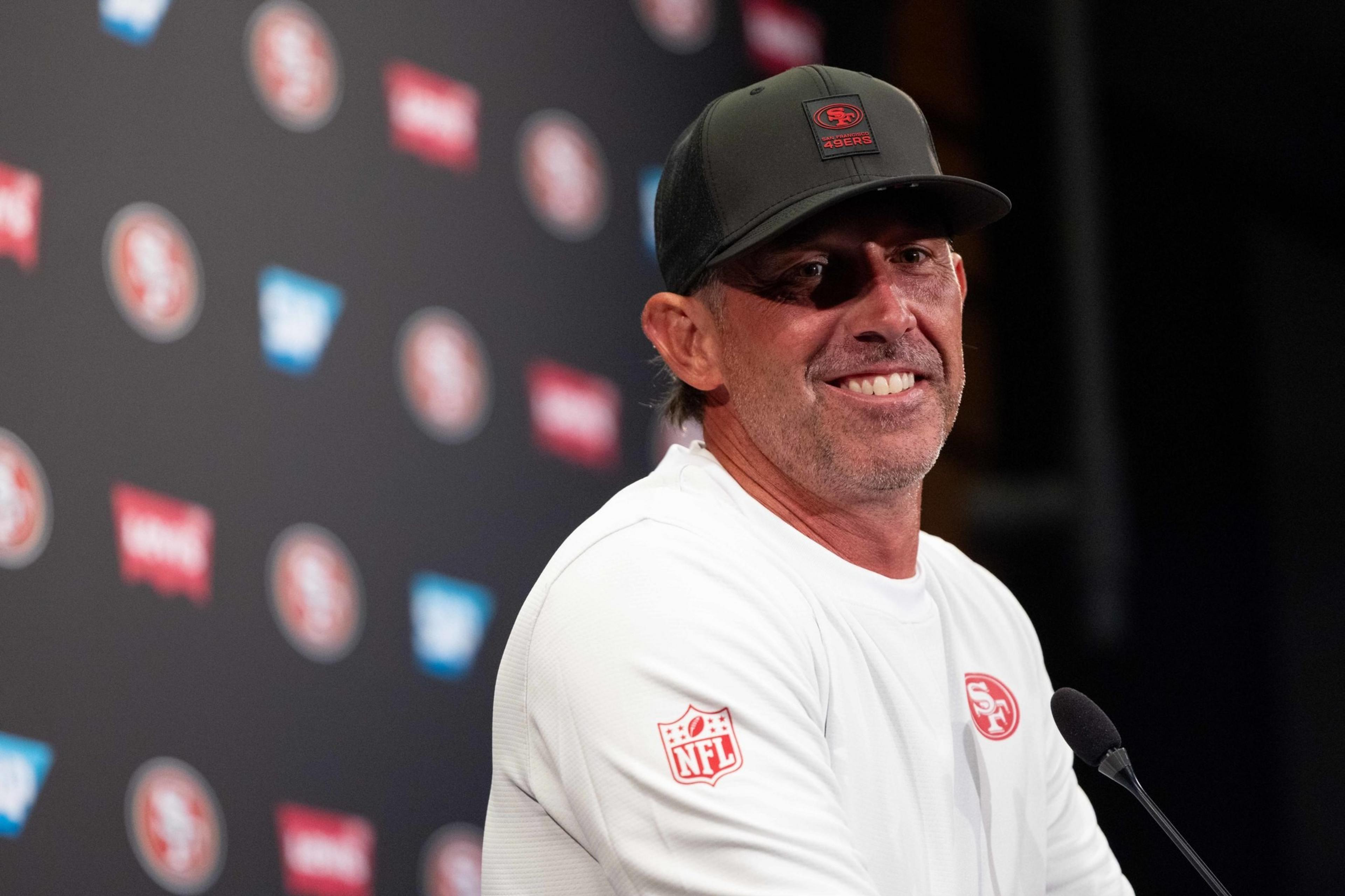 A man in a white sports shirt and black cap smiles at a microphone against a backdrop with red and white logos.