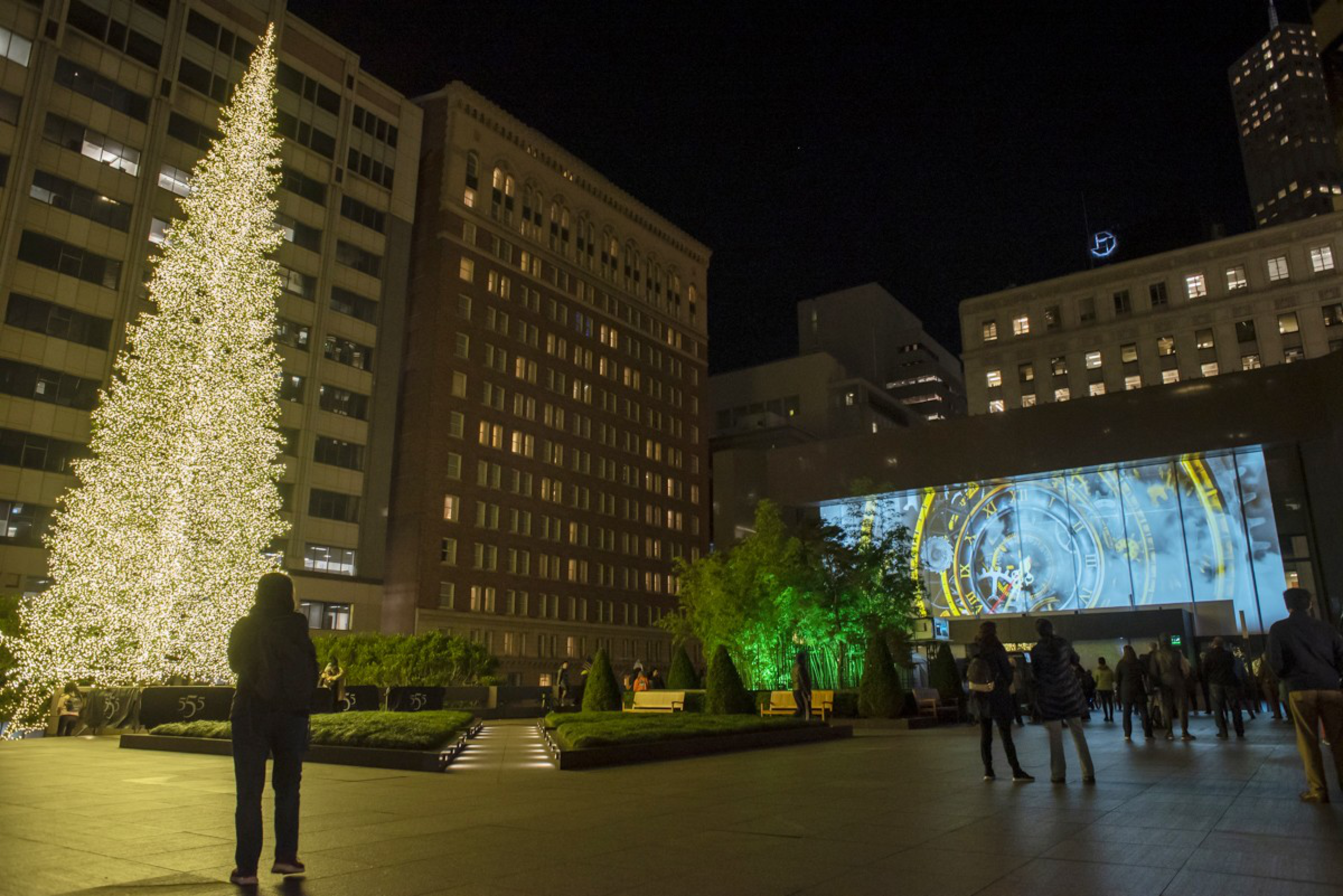 A large Christmas tree covered in white lights stands on the left, with people walking around a plaza surrounded by tall buildings at night.