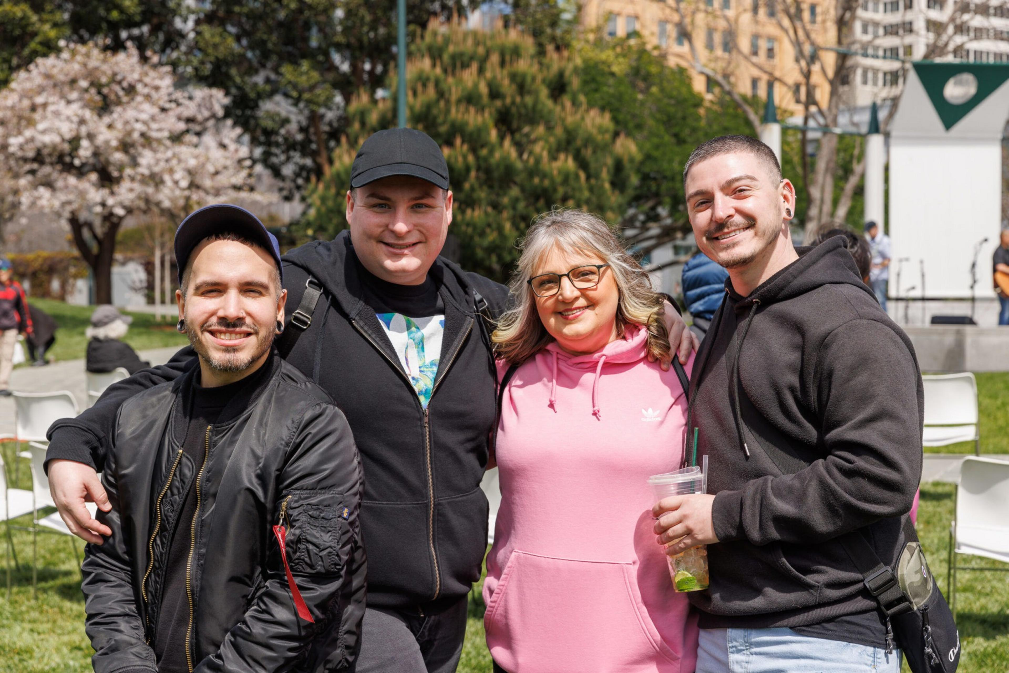 Four smiling people pose together in a sunny park with blooming trees and chairs in the background.