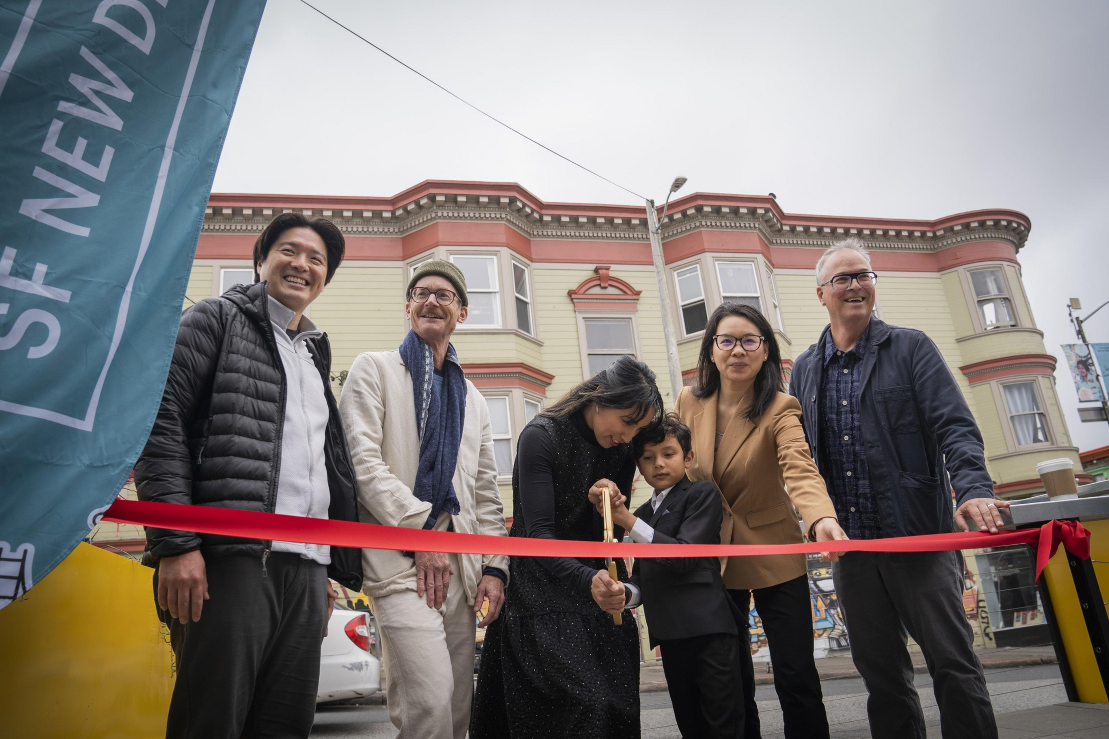 A group of six people, including a child, are smiling during a ribbon-cutting ceremony in front of a building. One person holds scissors with the child.