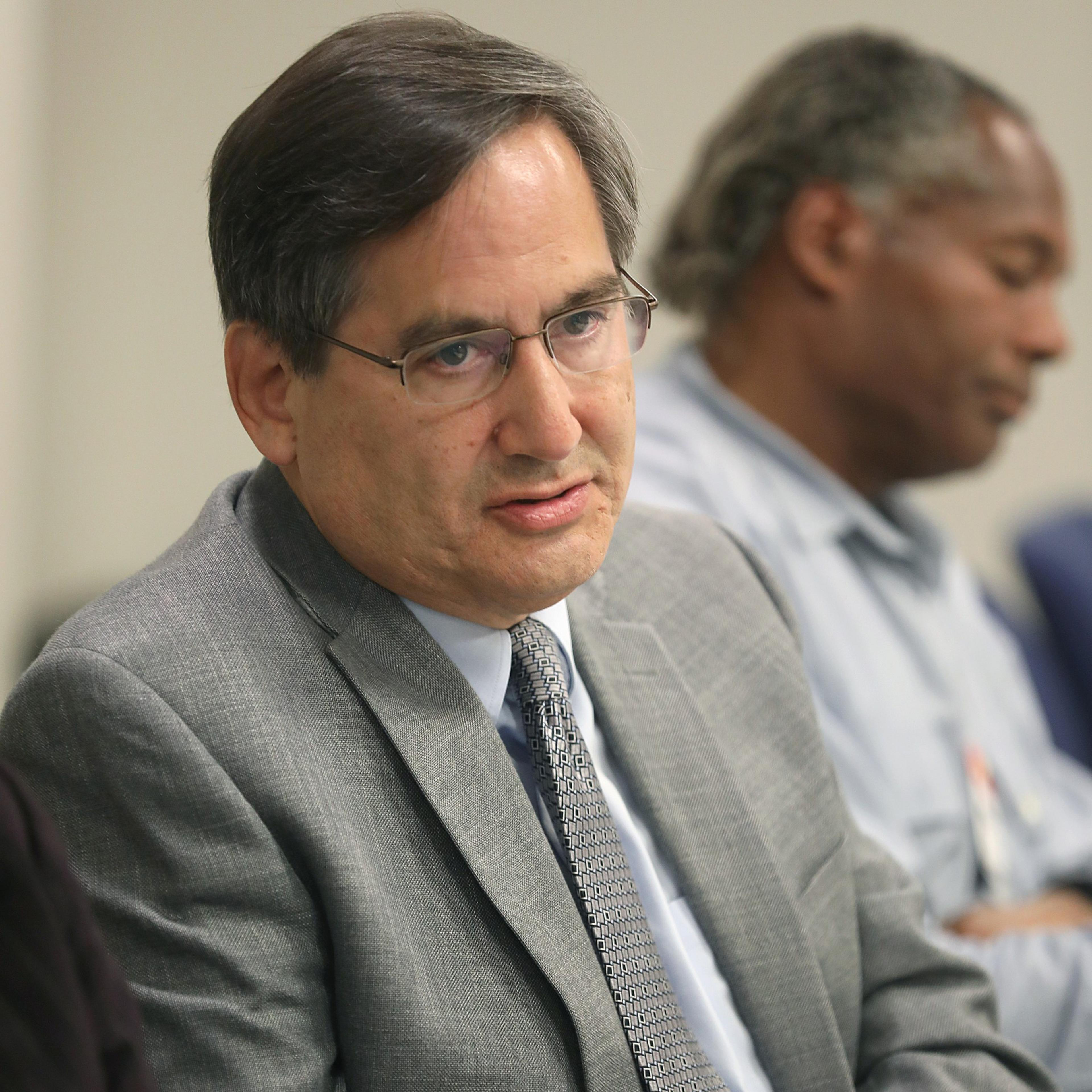 A man in suit and glasses looks in the direction of someone he's speaking with and a man behind me looks downward.