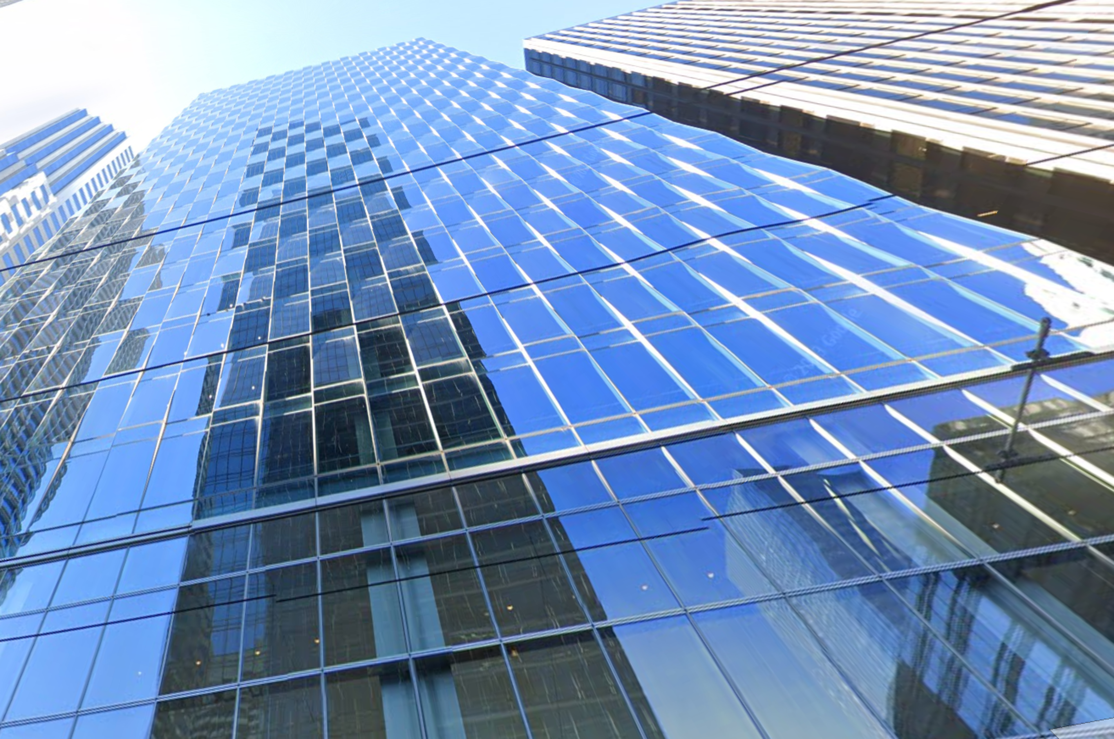 The image shows a tall, modern skyscraper with a reflective glass facade, glistening under a clear blue sky. Nearby buildings are also reflected on its surface.