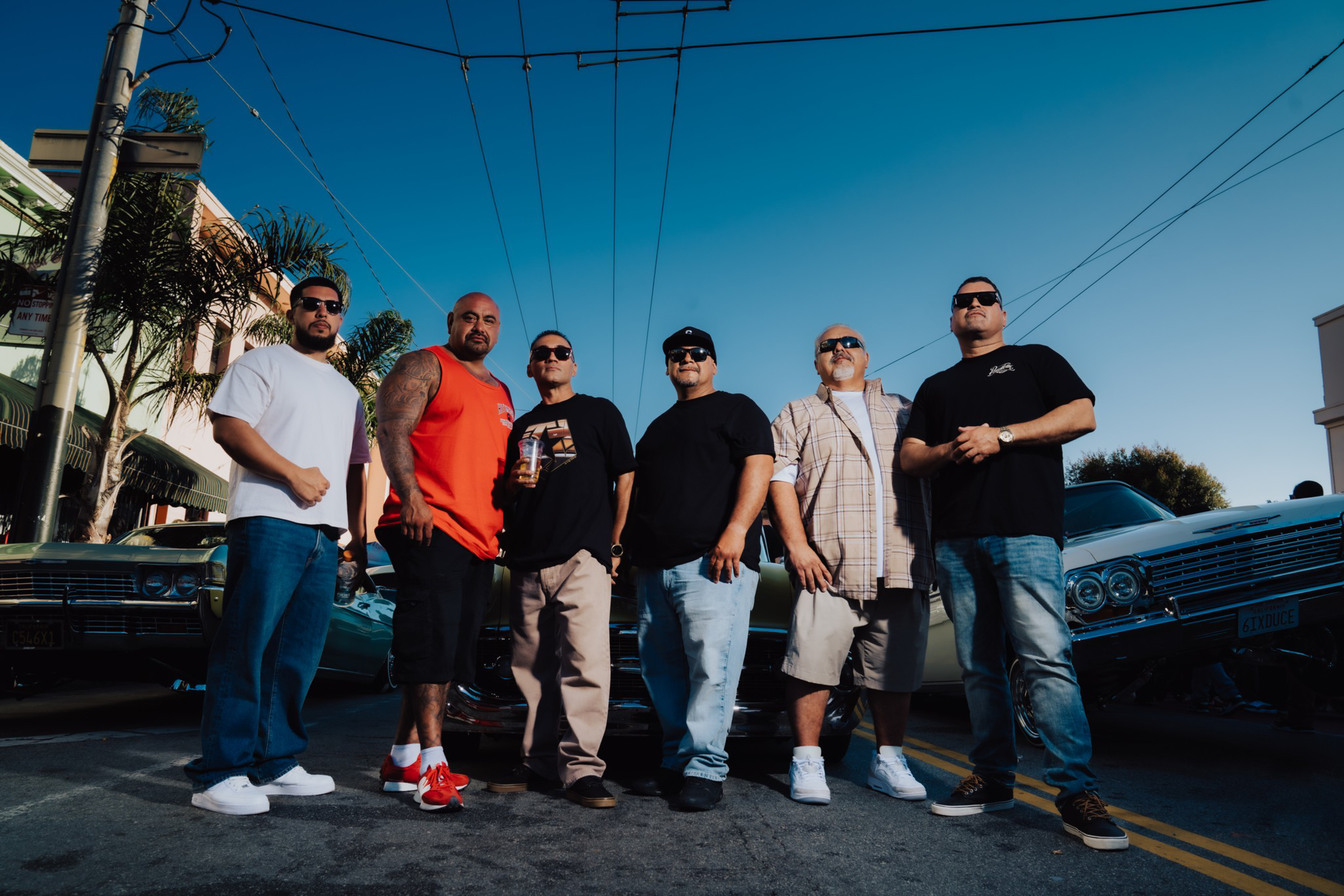 Six men stand confidently in a street flanked by vintage lowrider cars, wearing casual streetwear and sunglasses under a clear blue sky.