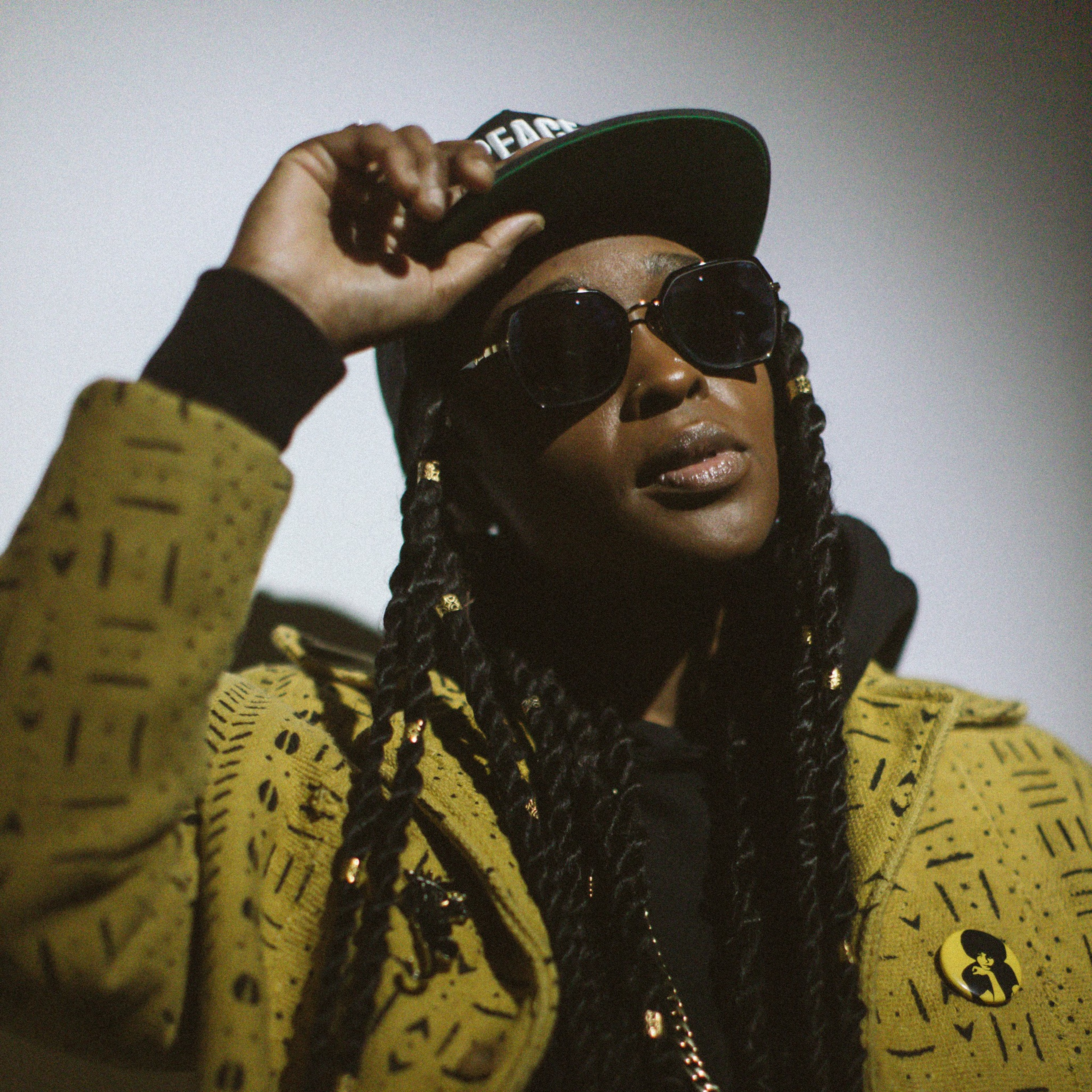A person with long braided hair, wearing large sunglasses, a black cap, and a patterned mustard yellow jacket, poses with one hand adjusting the cap.