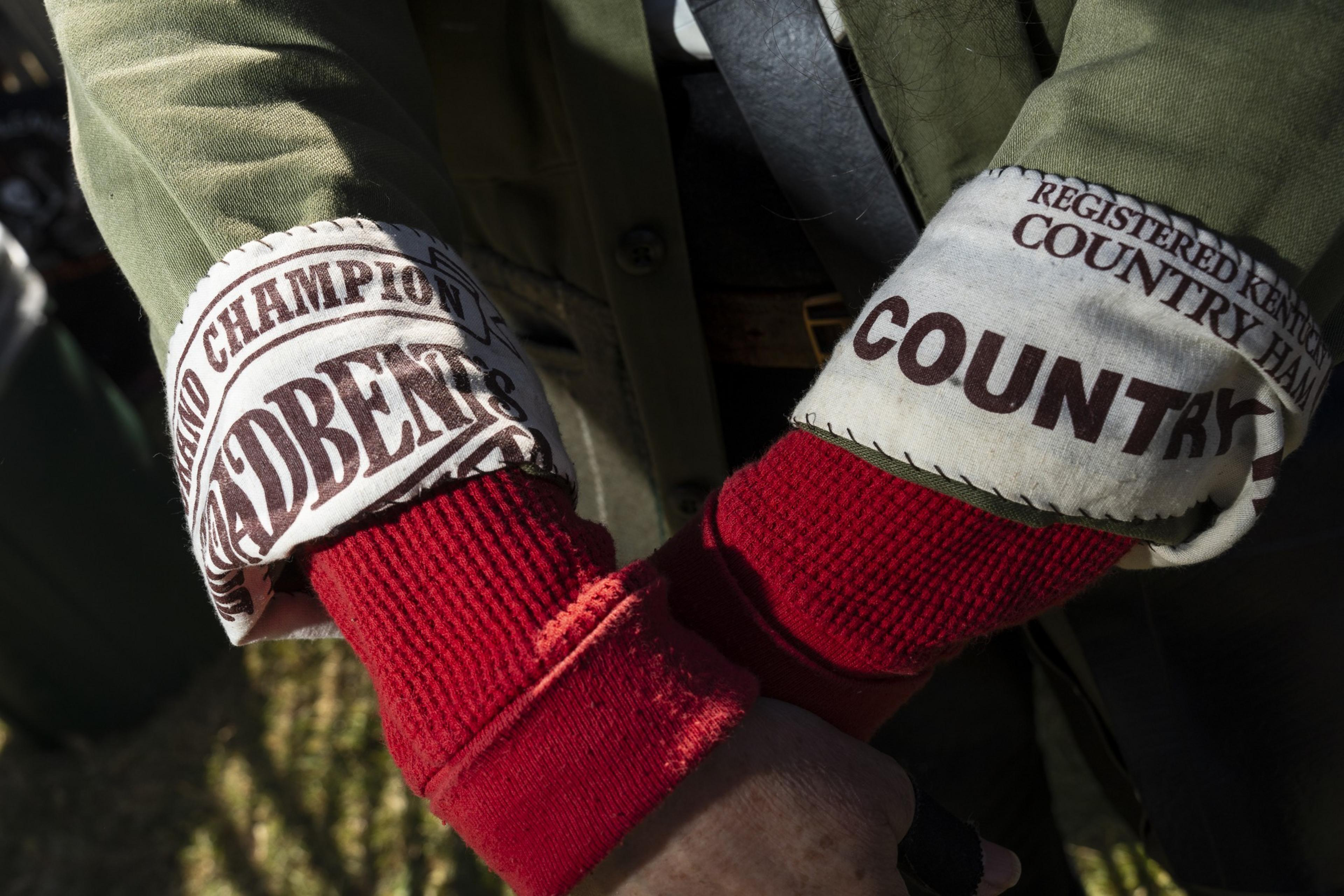 The image shows a person wearing a green jacket with the sleeves rolled up, revealing red thermal sleeves beneath. The cuffs have printed text about country ham.