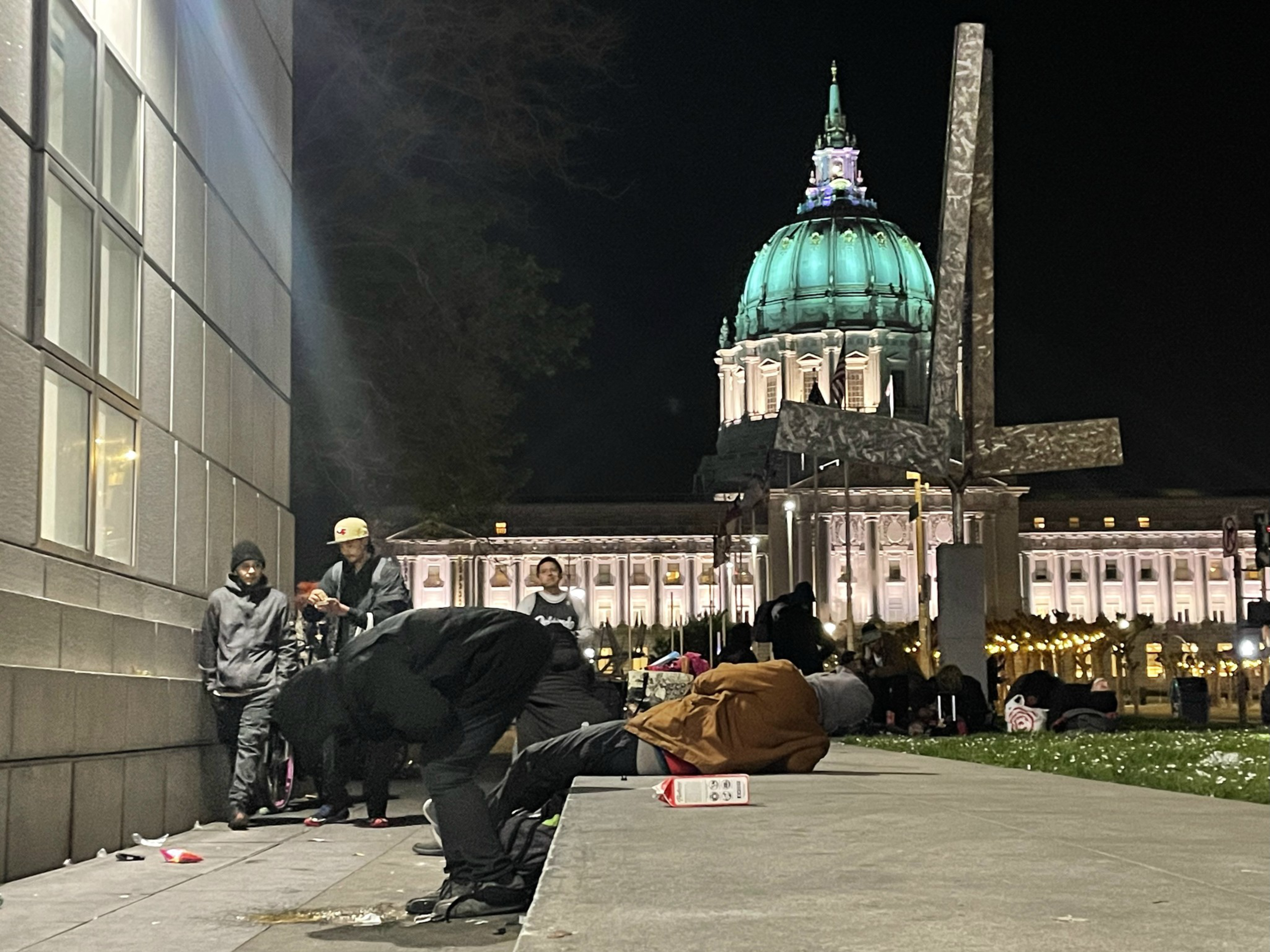 A group of people gathers at night near a lit-up domed building, with a large abstract sculpture in the foreground.