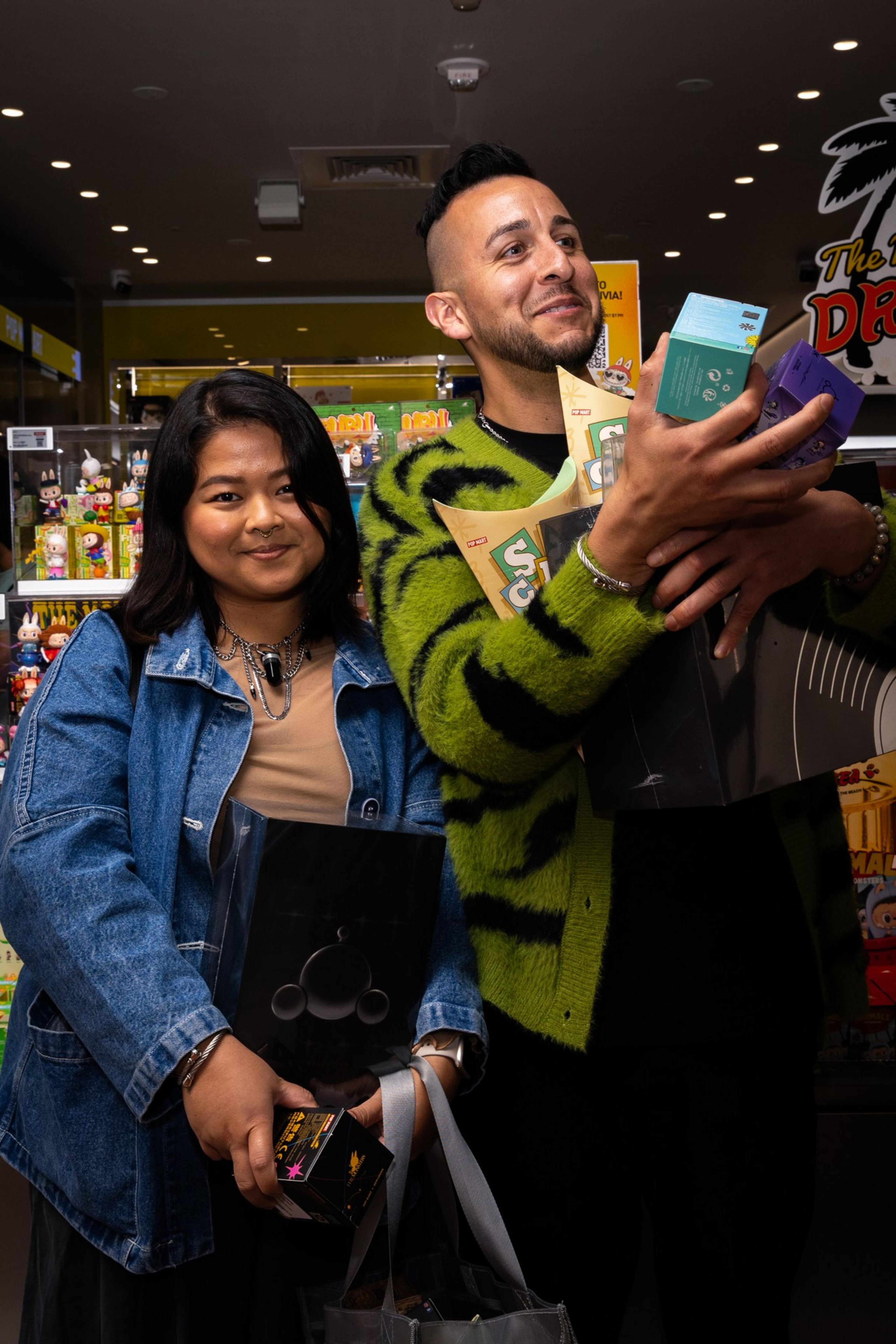 A smiling woman in a denim jacket and a man in a green patterned sweater hold multiple items in a store filled with colorful toy displays.