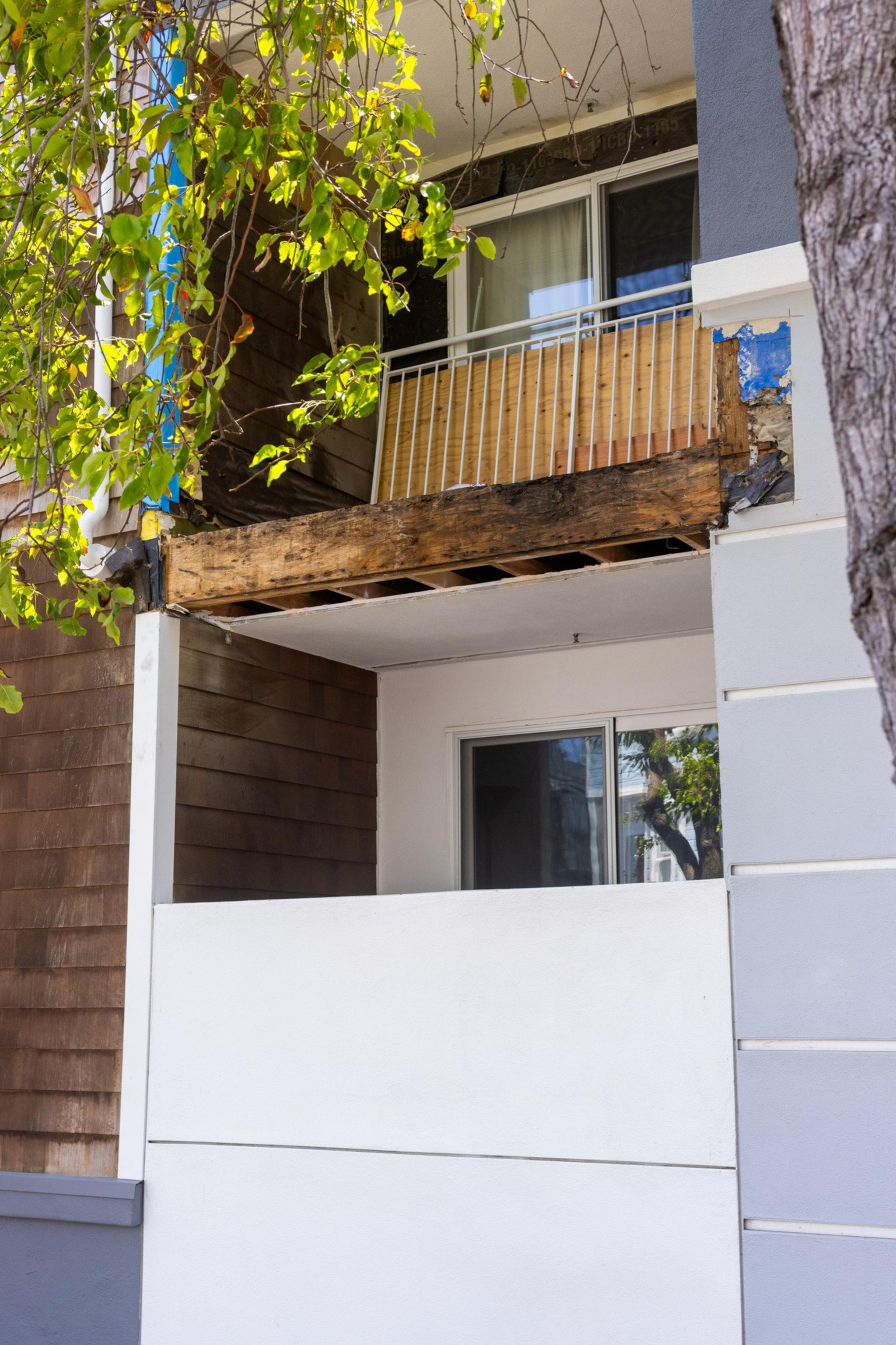 The image shows a damaged balcony with missing panels, blue tarp, and exposed wooden beams. A tree with green leaves partially obscures the view.