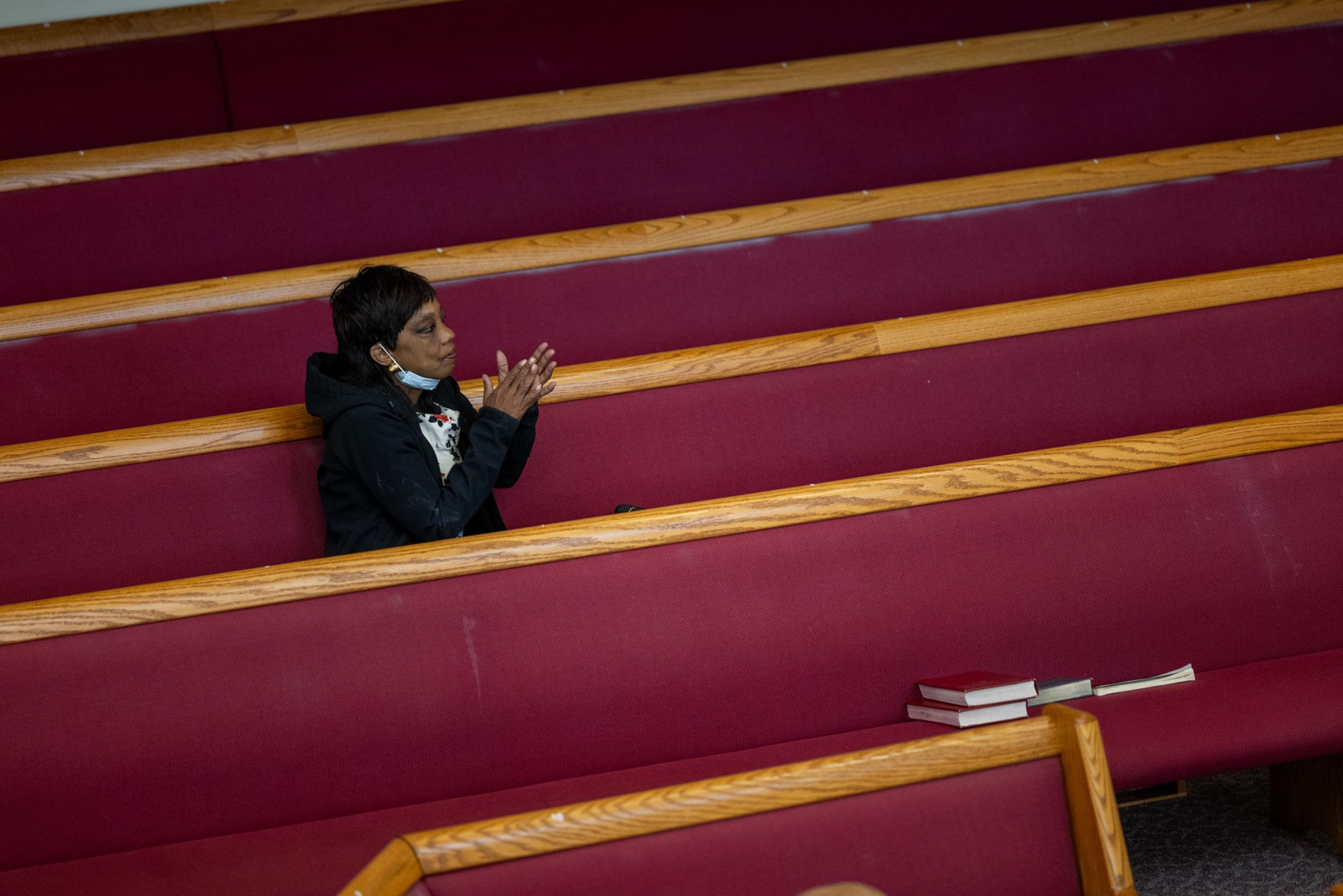 A person sits alone in a red church pew, clasping their hands in prayer. There are a few stacked books at the other end of the pew.