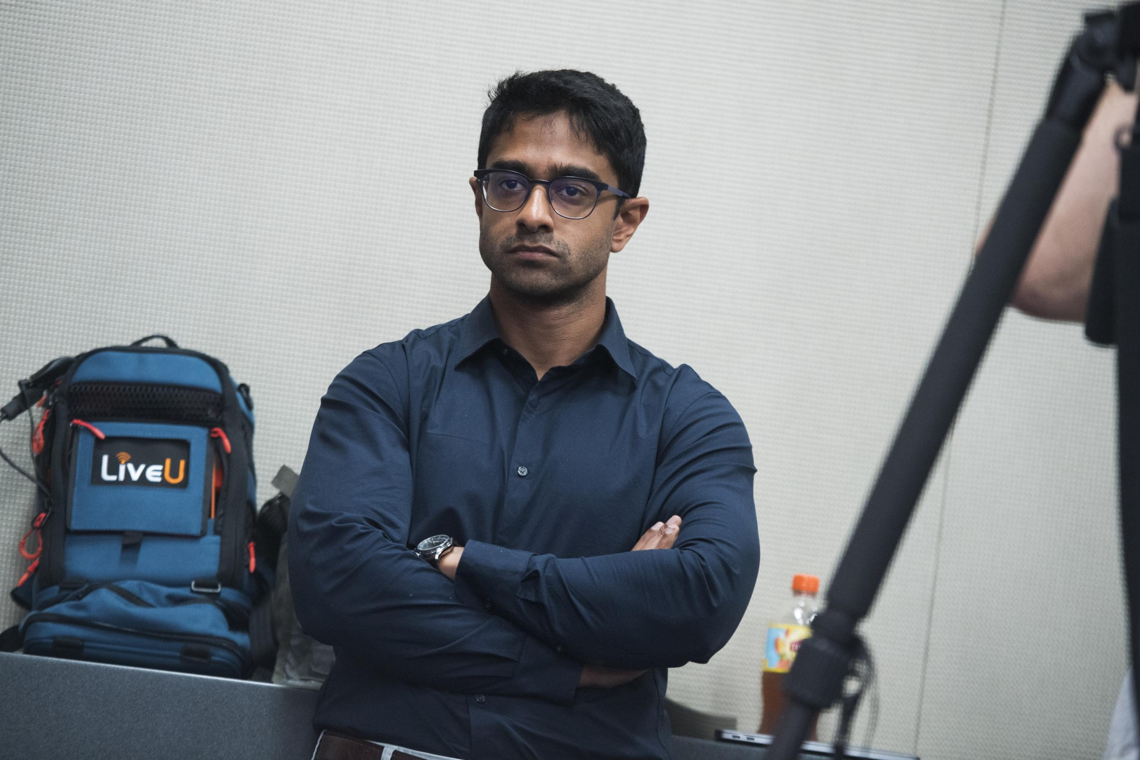 A person with glasses and a dark shirt stands with crossed arms. A blue backpack with "LiveU" is nearby, and a bottle with an orange cap is on the table.