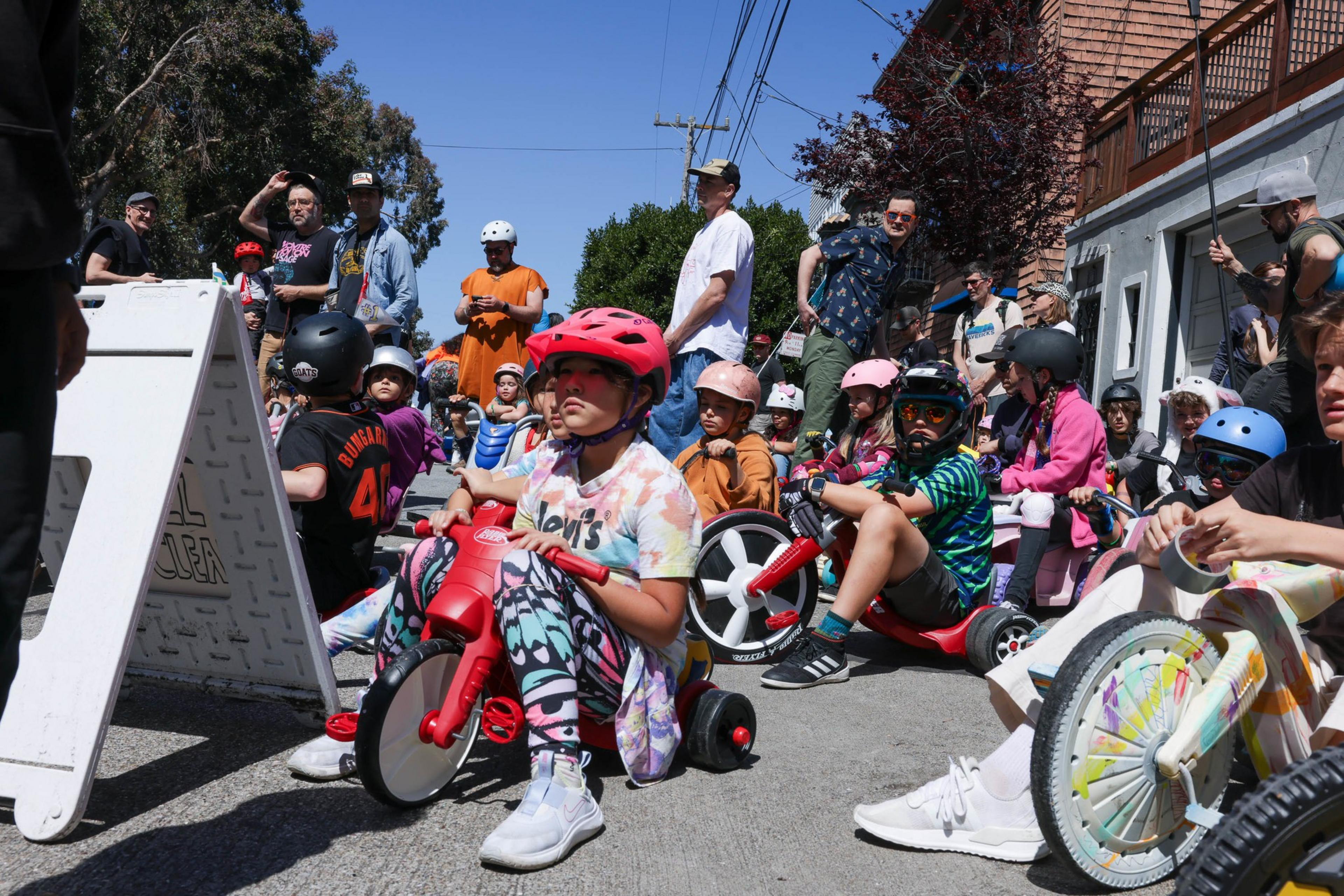 Children on toy cars at a starting line, with spectators in a sunny outdoor setting.