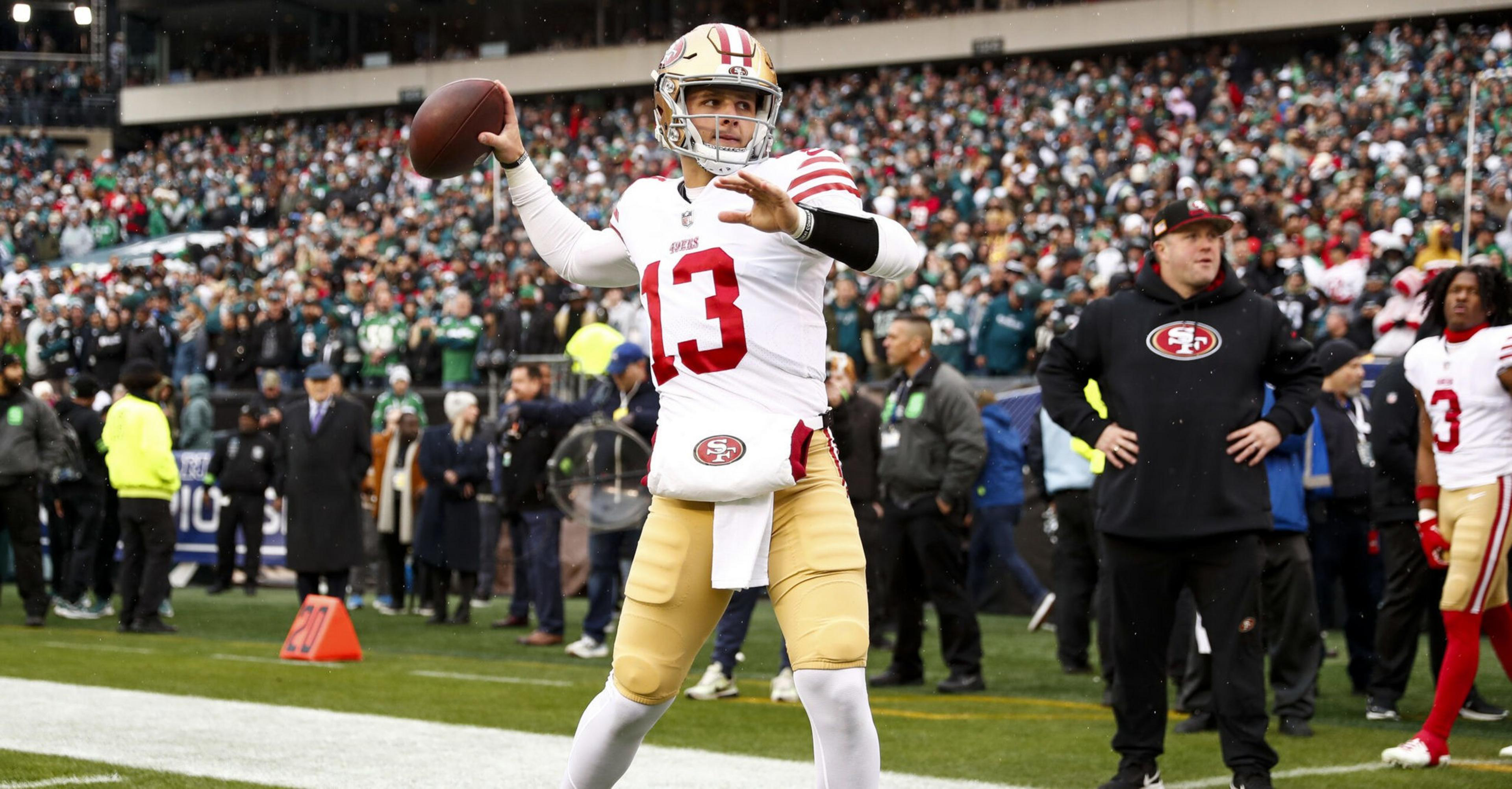 A football player in a white and red San Francisco 49ers uniform prepares to throw a pass on the sideline, with coaches and fans in the background.