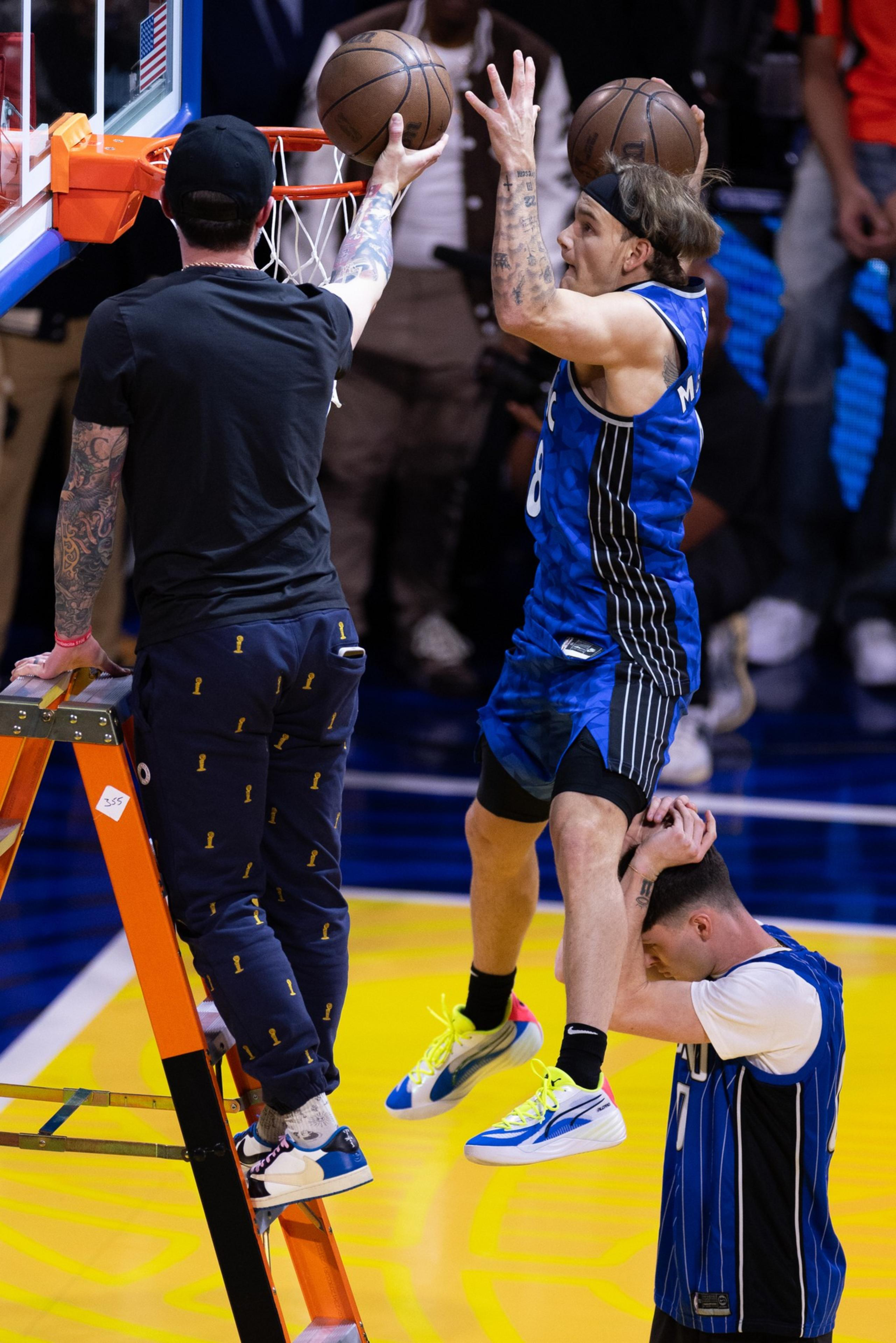 A basketball player in a blue jersey jumps, supported by a teammate holding his legs. He is reaching to dunk a ball, while another person stands on a ladder.