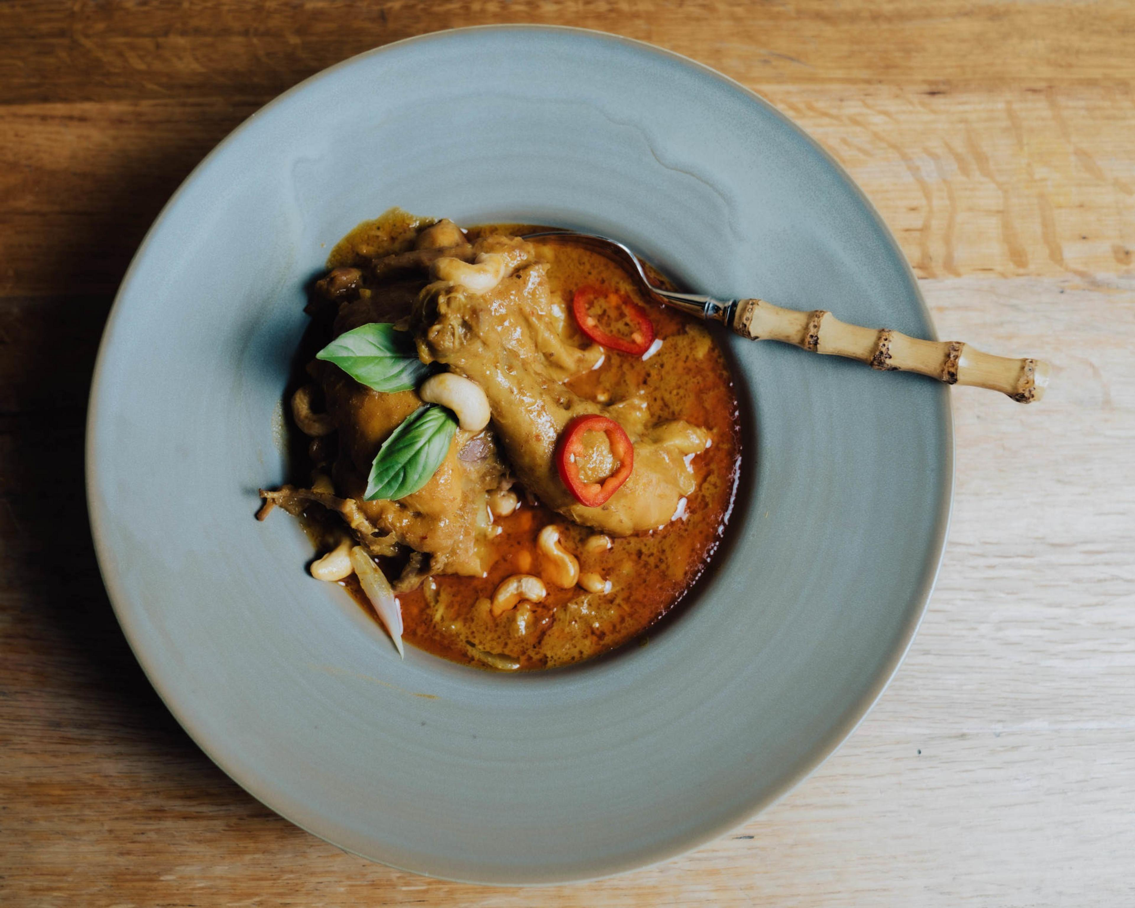 Chicken drumsticks in a creamy brown curry sauce, garnished with basil leaves, red chili slices, and cashew nuts, served in a blue bowl with a spoon.