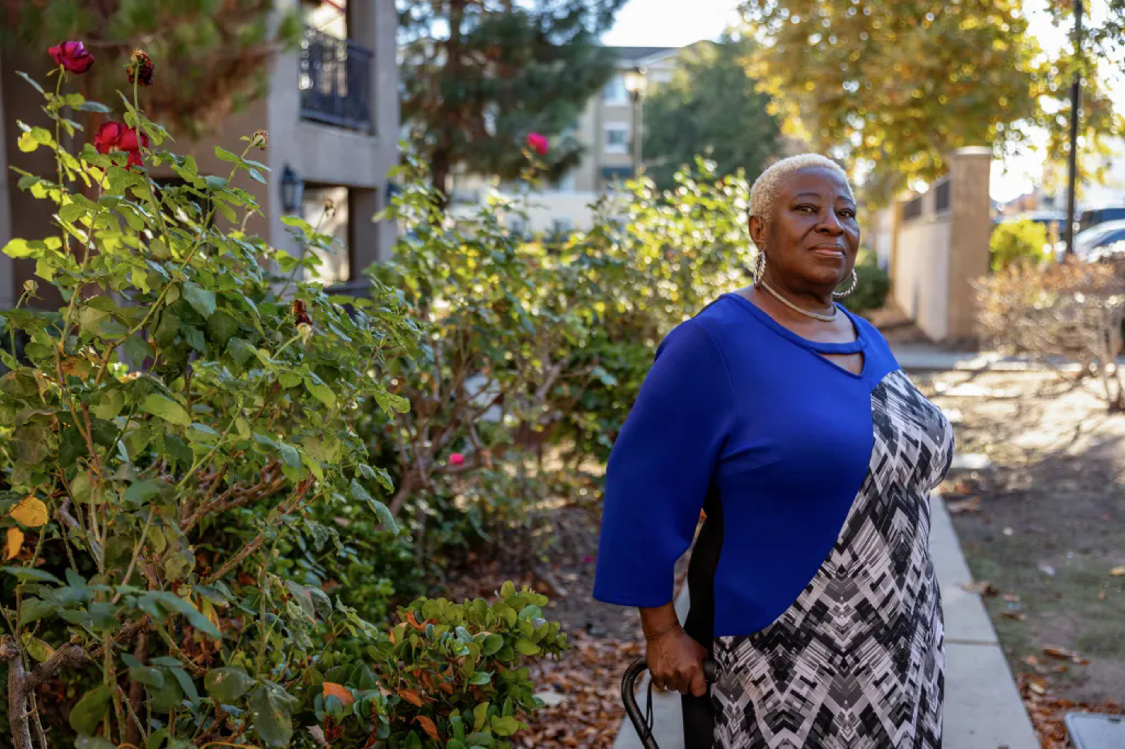 Woman in blue, black and white dress stands with cane next to rose bushes outside apartment building