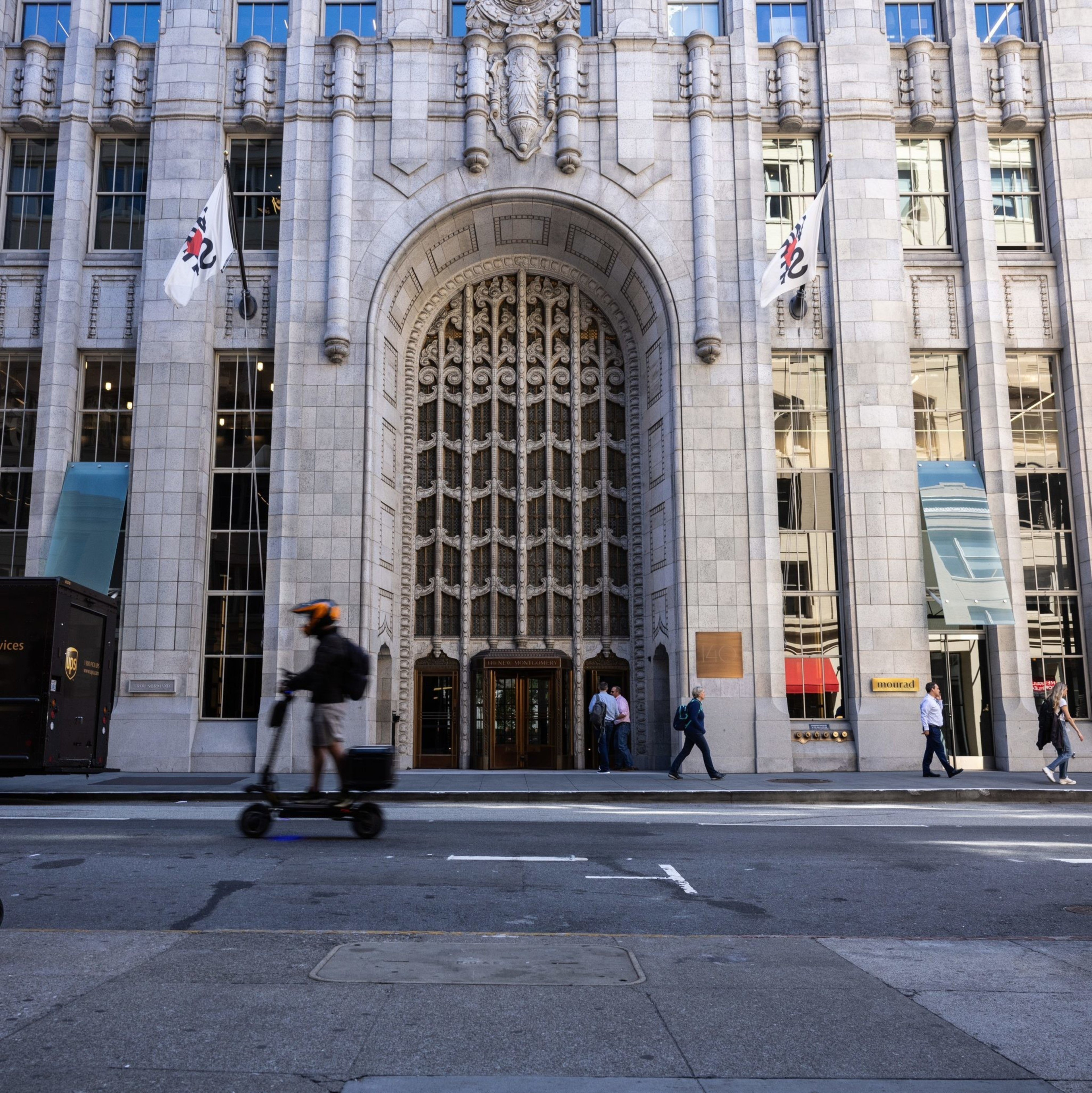 A large building with ornate architecture and tall windows has a grand arched entrance. People walk and ride scooters in front, while a UPS truck is parked nearby.