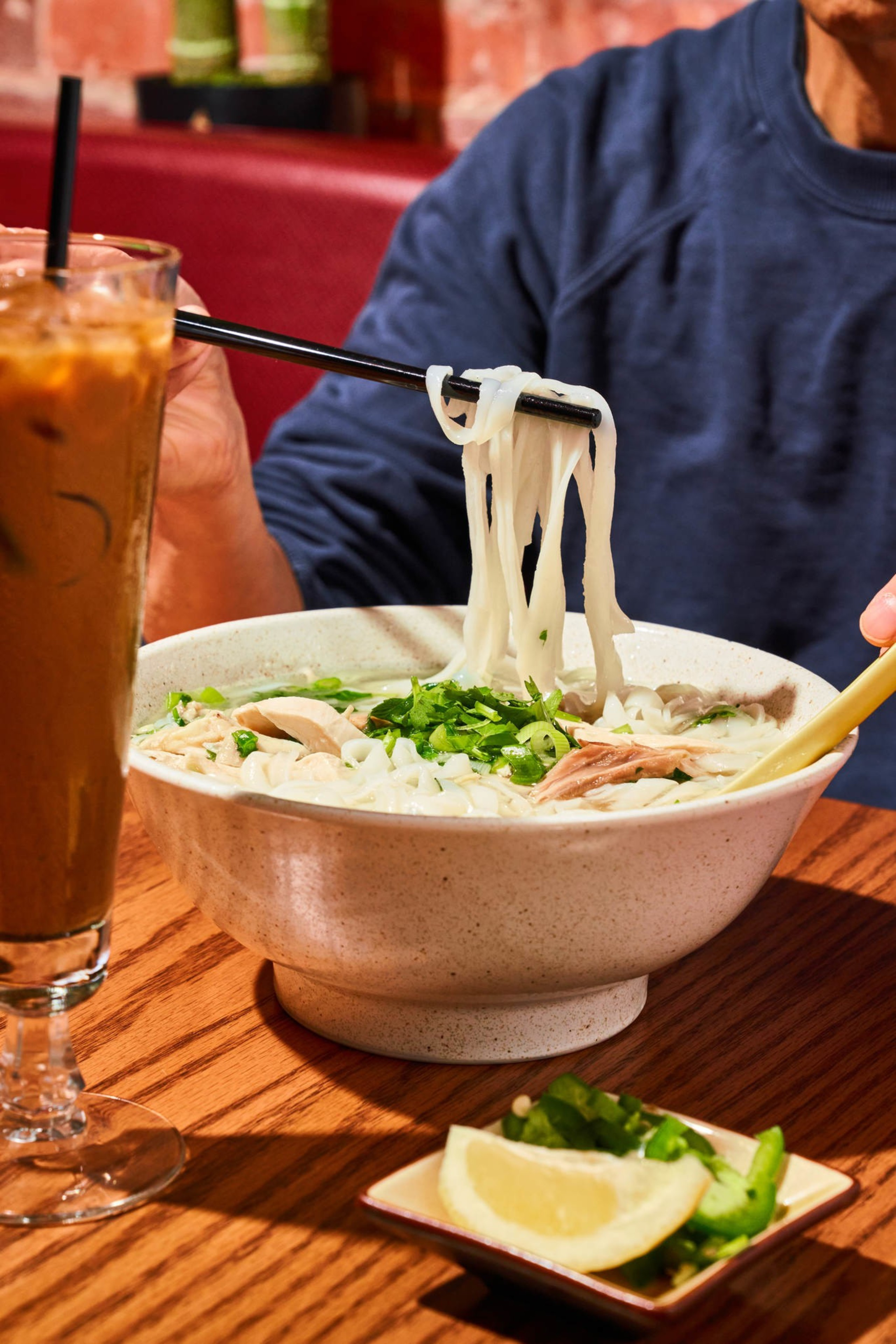 A person in a blue shirt holds chopsticks lifting noodles from a large bowl of pho topped with herbs. An iced drink and lemon slices are nearby.