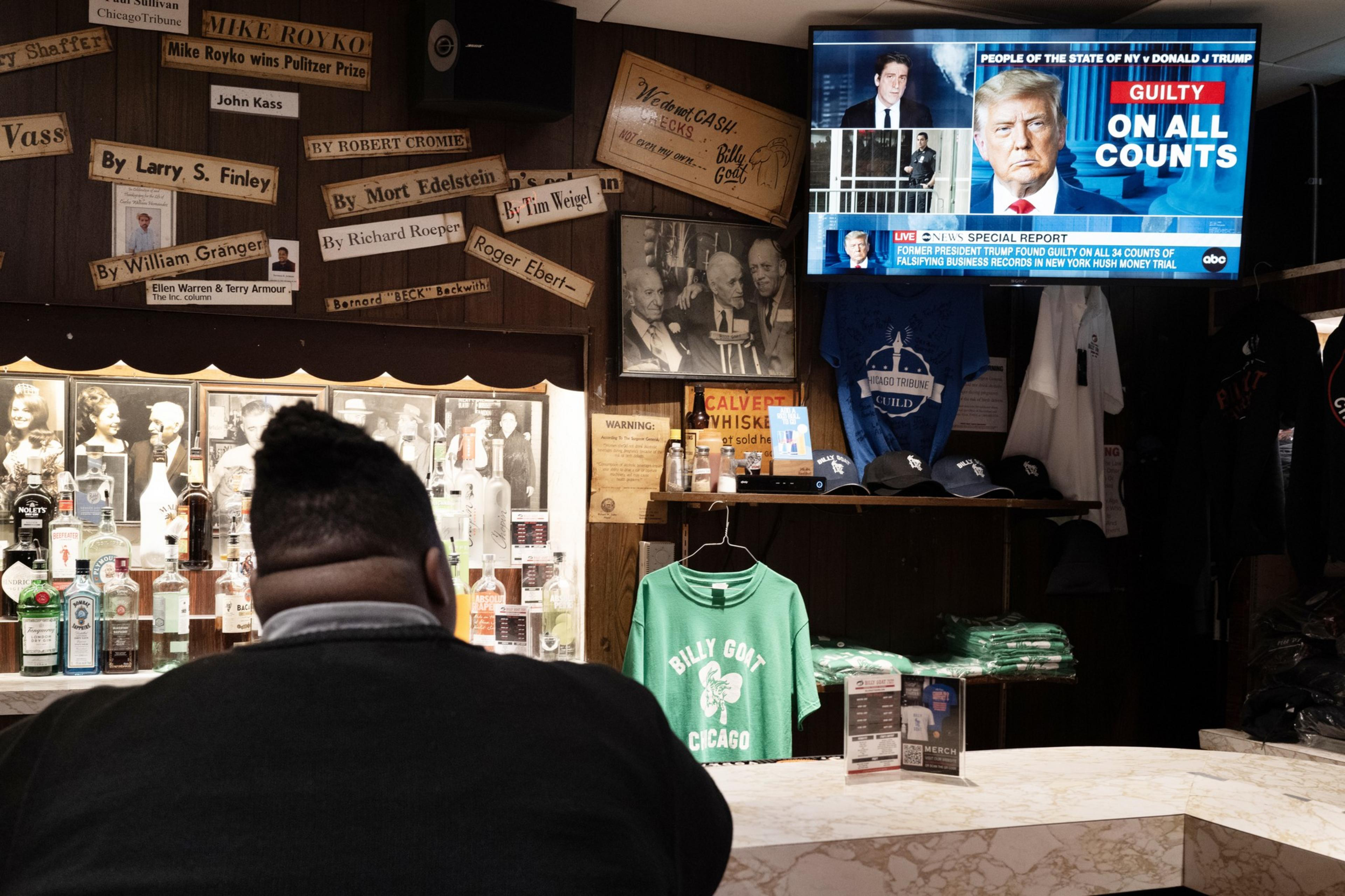 A person sits at a bar viewing a TV that displays a headline saying "Guilty on all counts" with a picture of a man. Bar shelves have liquor bottles, merchandise, and framed photos.