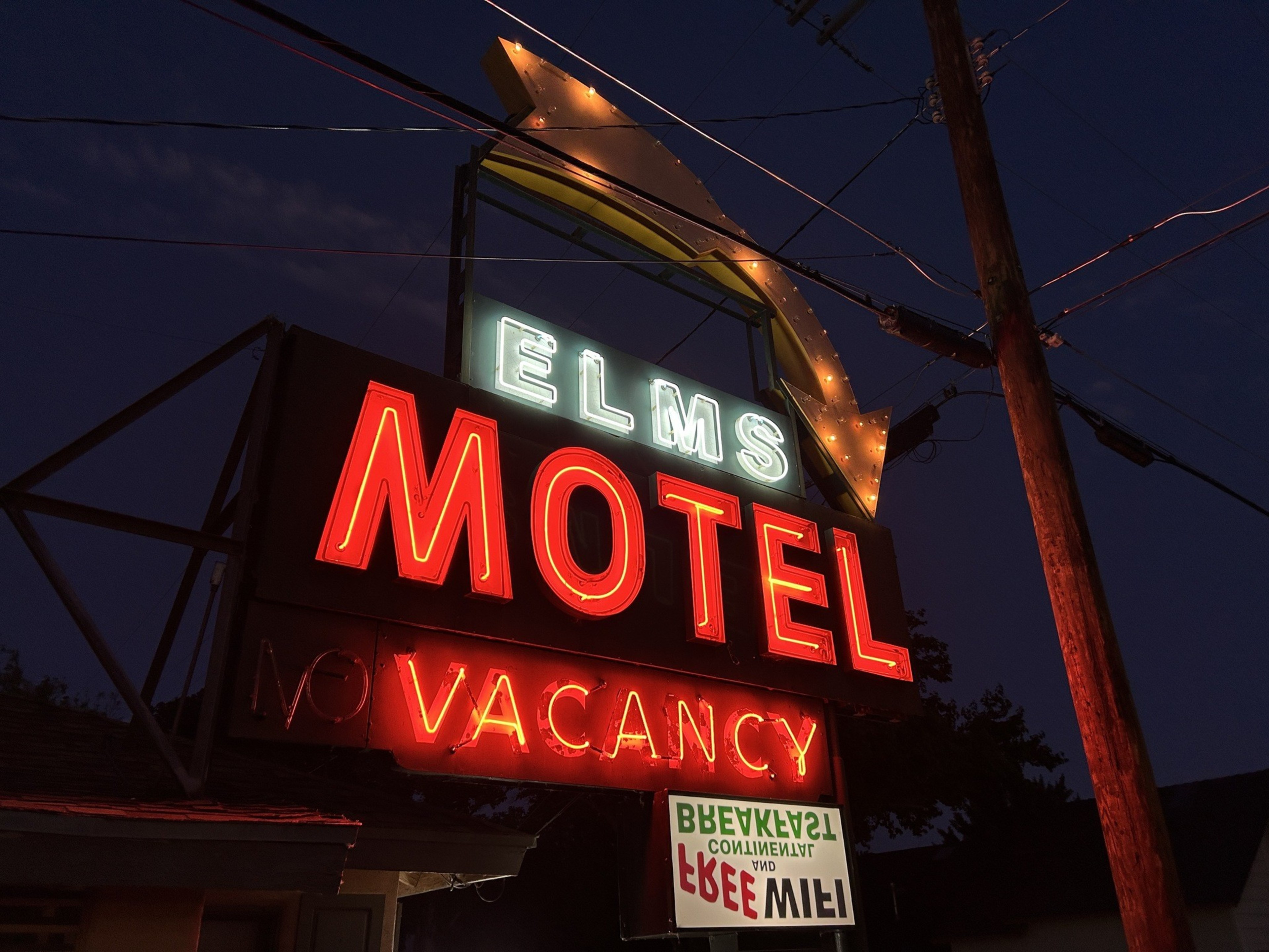 A brightly-lit neon sign reads &quot;Elms Motel.&quot; The word &quot;Vacancy&quot; is illuminated in red beneath the main sign, and a lit arrow points downwards.