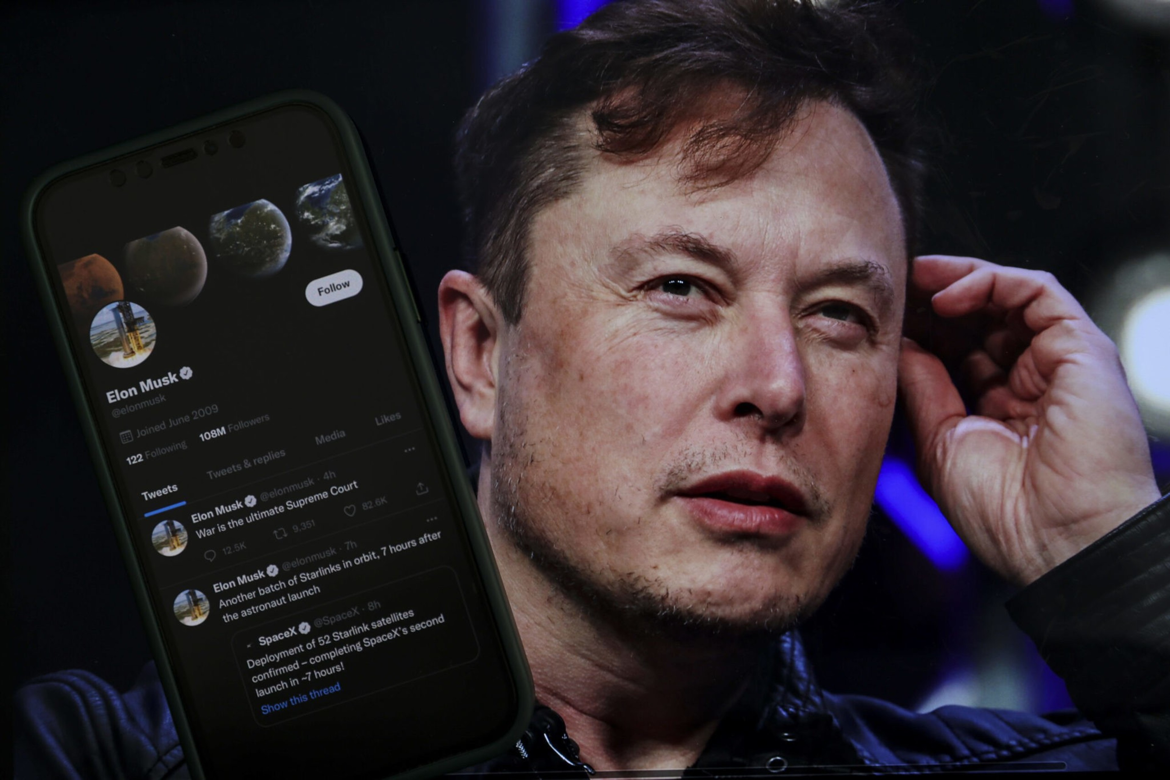 A man is touching his ear, looking thoughtful. In front of him, there's a smartphone displaying a Twitter profile with the name "Elon Musk" and 108M followers.
