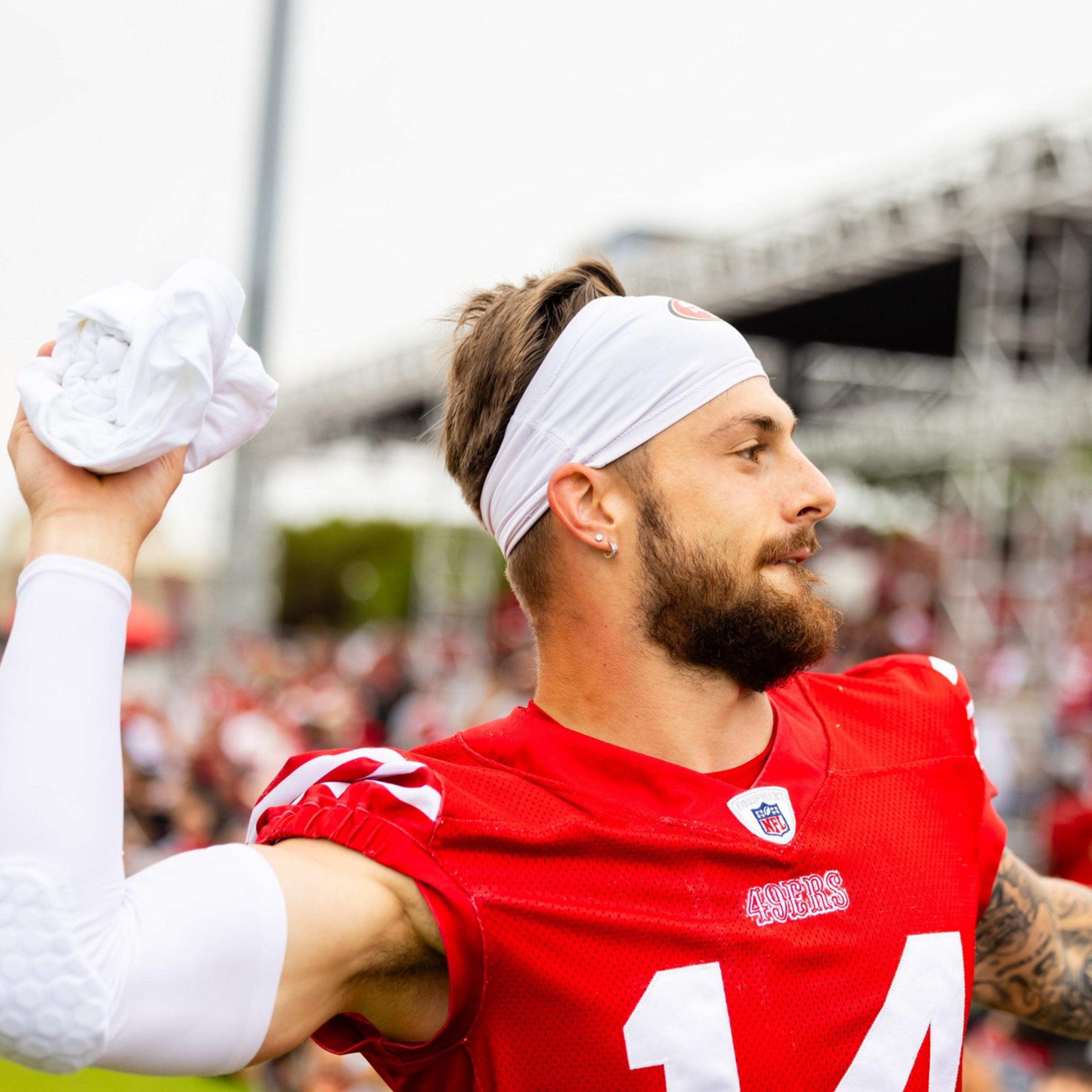 A football player in a red jersey, wearing a white headband and arm sleeve, throws a towel toward the audience at what appears to be a training event or game.