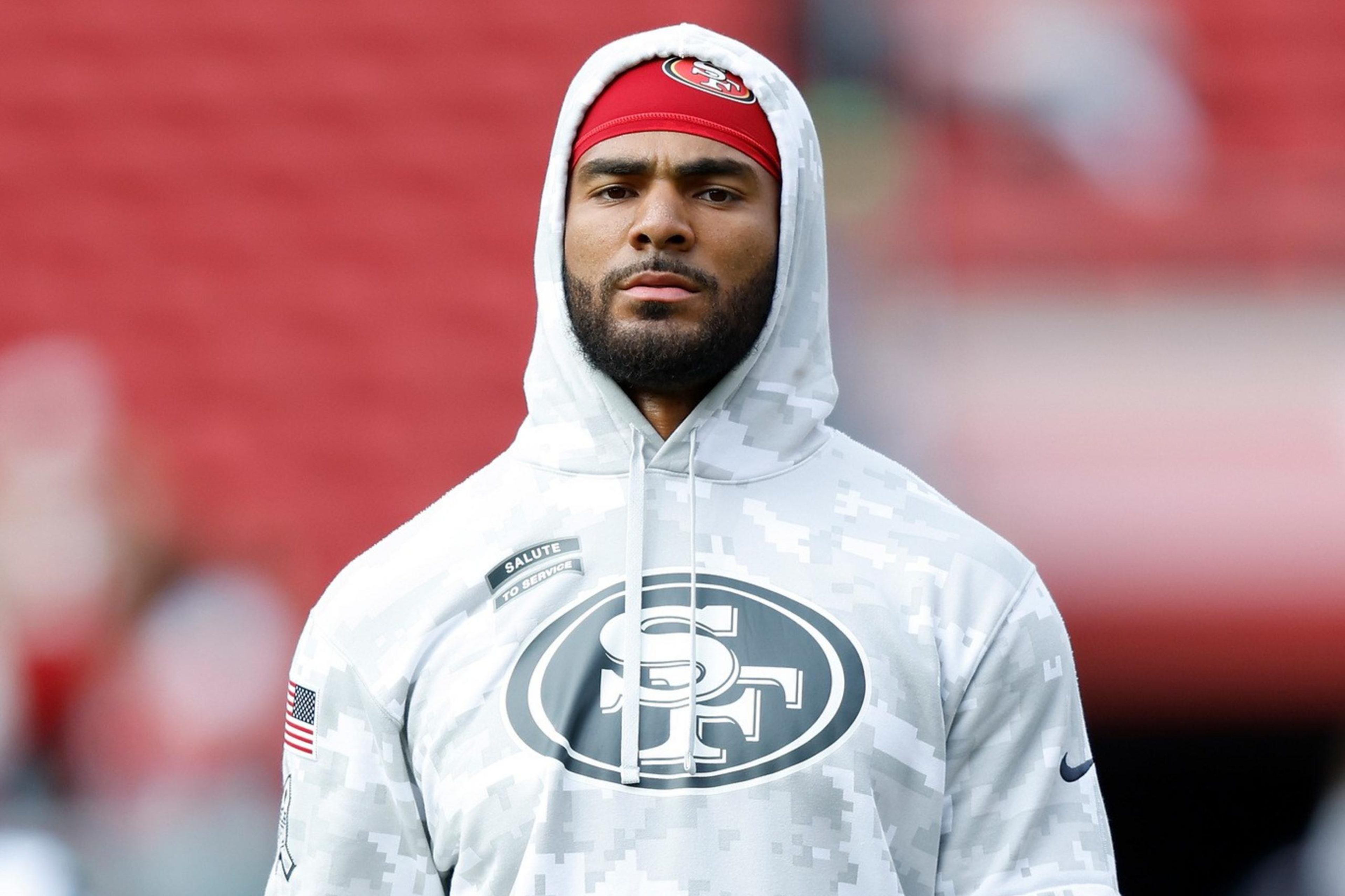 A man in a camouflage hoodie with an "SF" logo holds a football in a stadium. He wears a red cap, white gloves, and black shorts.