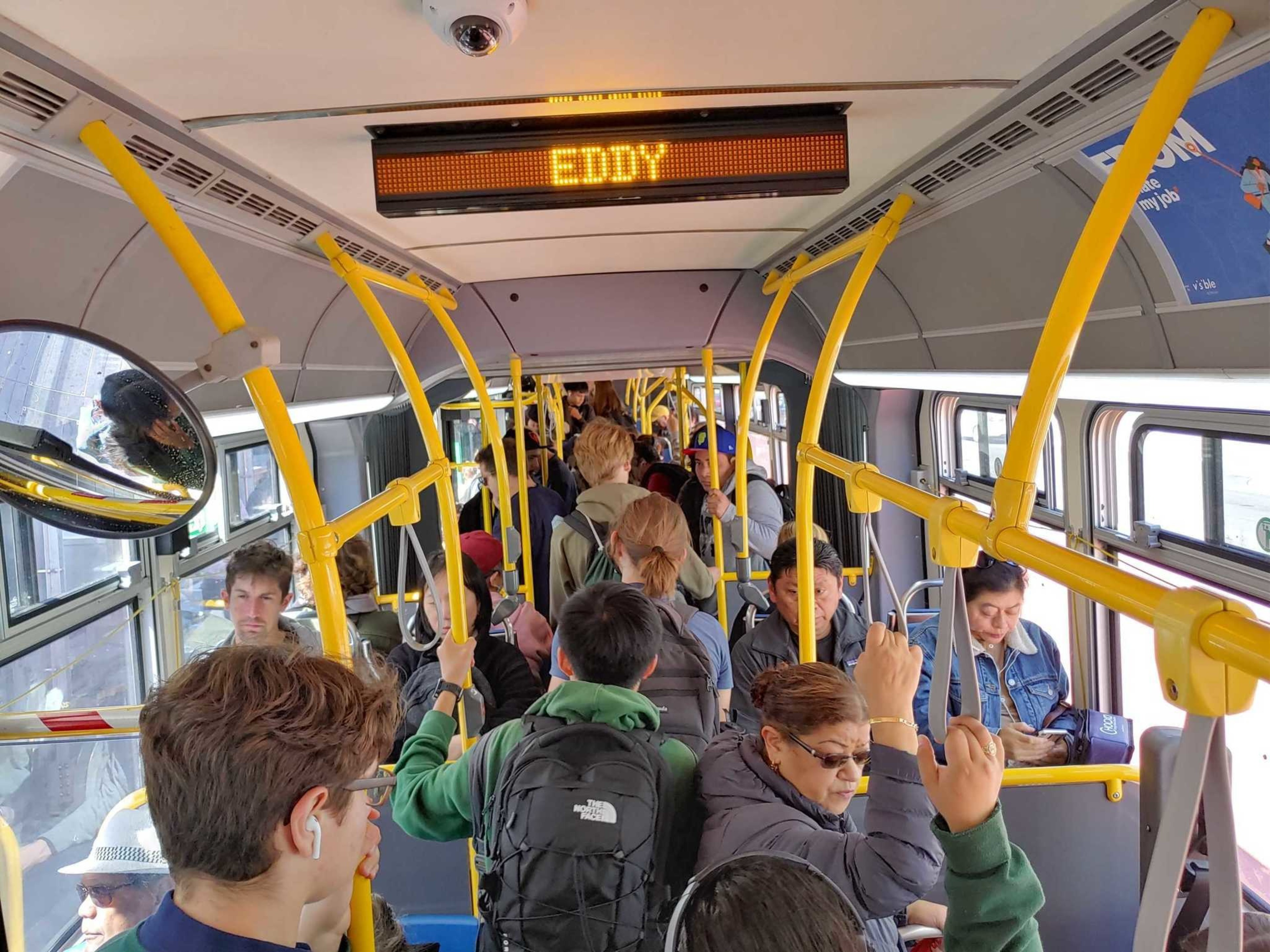 A crowded bus interior with passengers standing and sitting, yellow handrails, and an electronic sign displaying "EDDY."