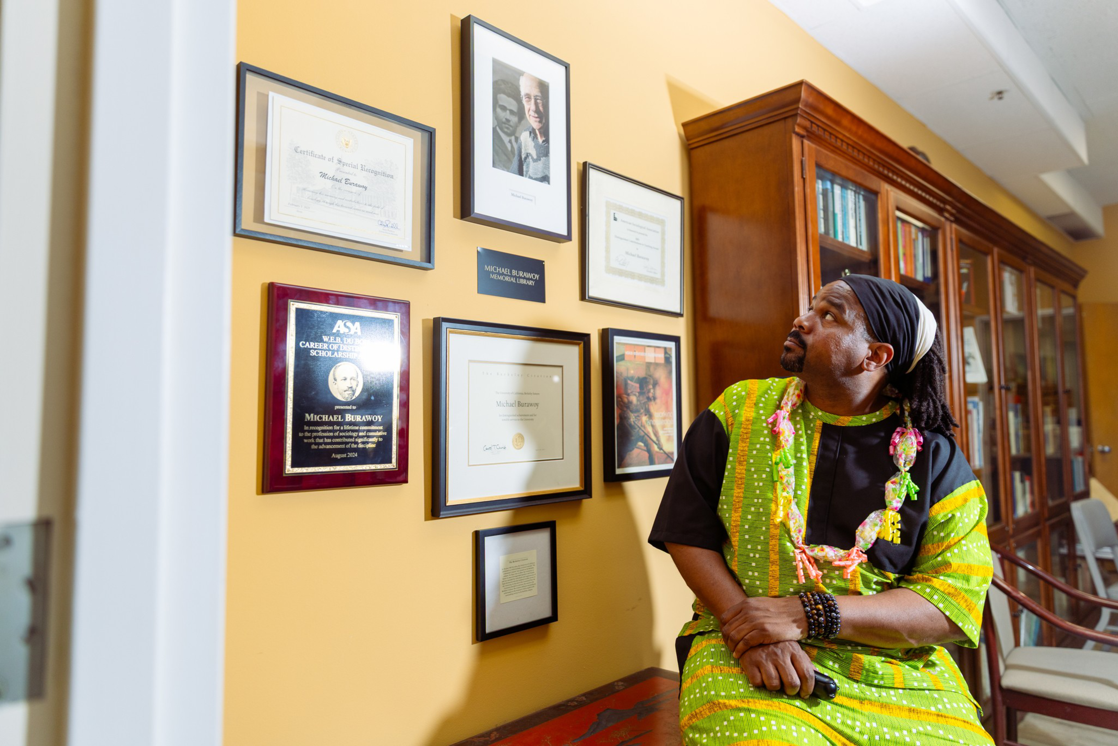 A man in a green and yellow patterned outfit sits on a bench, looking up at a wall displaying framed certificates and photos.