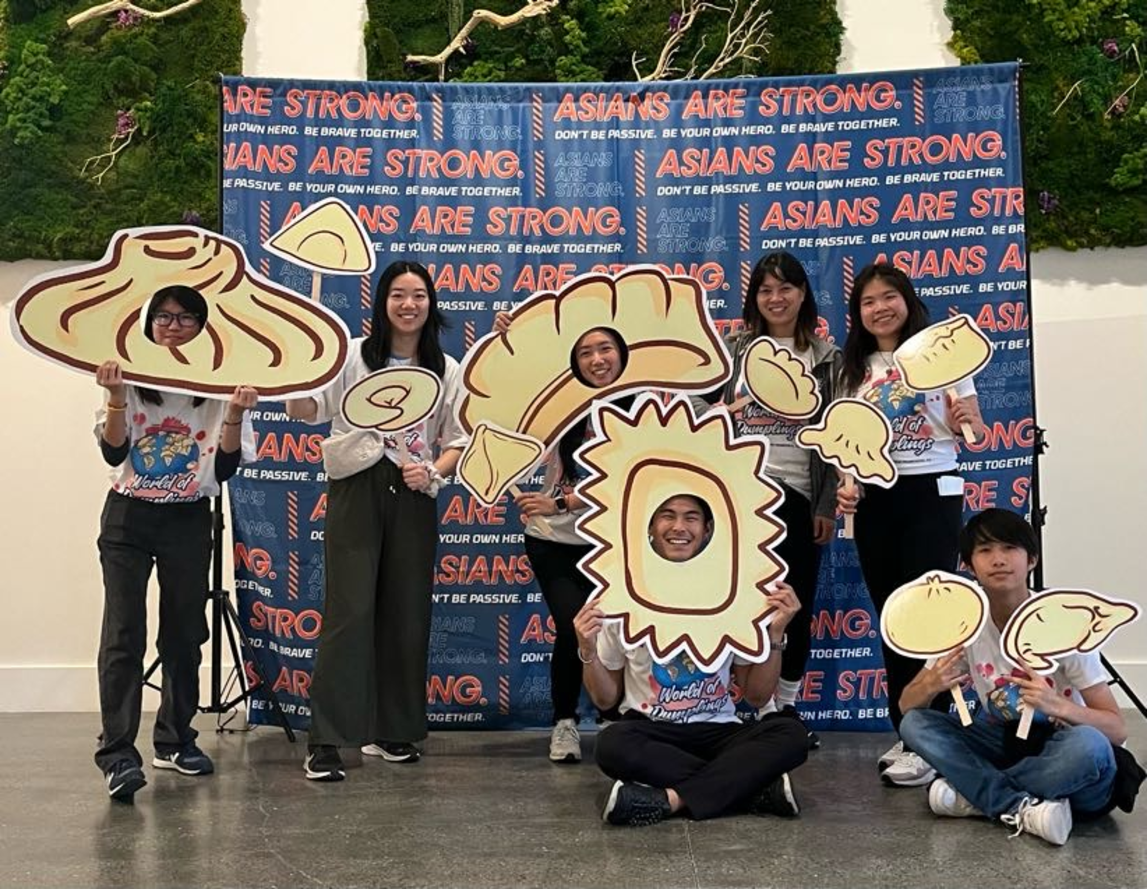 Seven people hold large cutouts of various dumplings, smiling in front of a backdrop that reads “Asians Are Strong.”
