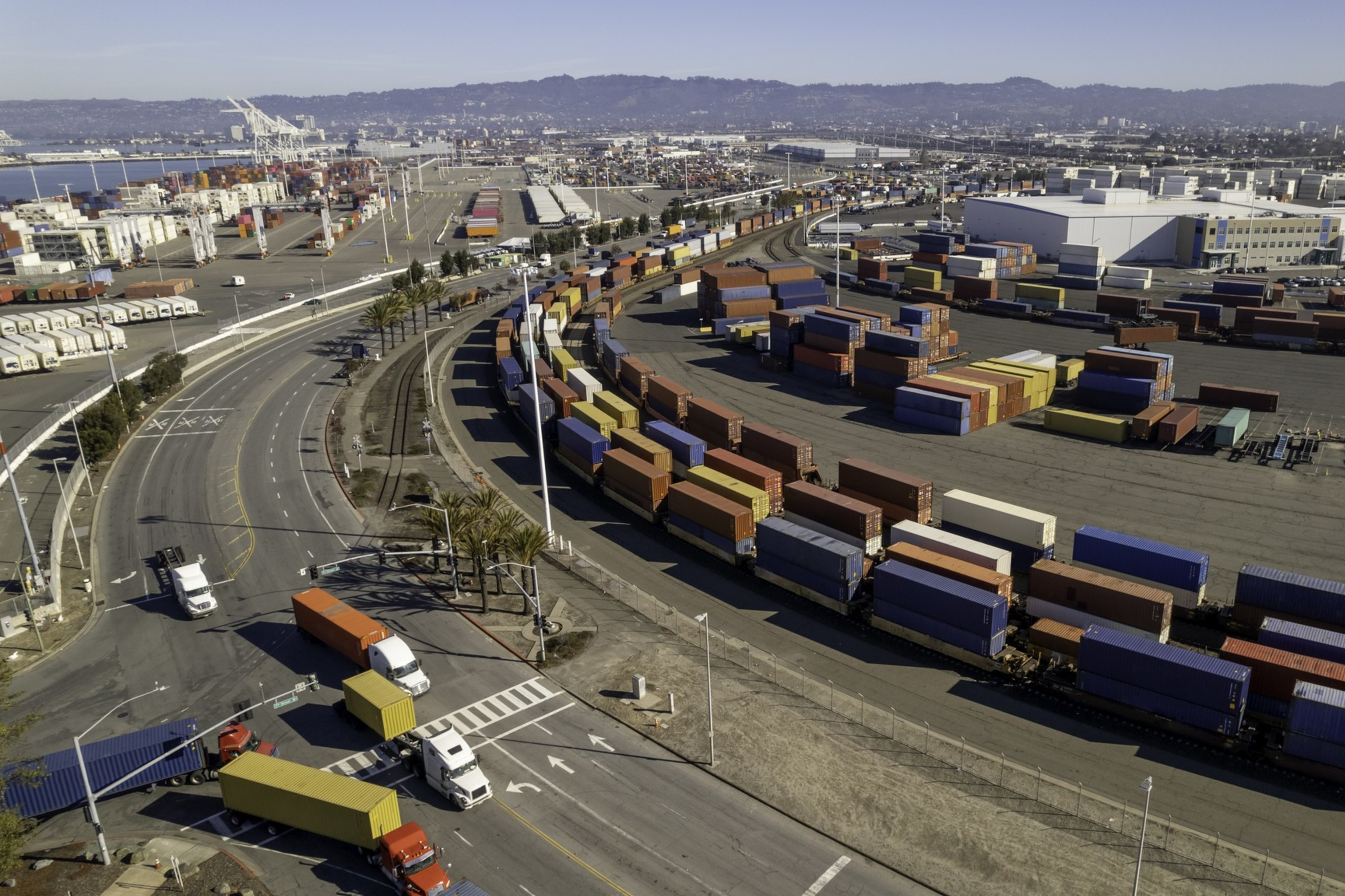 The image shows a large container yard with rows of colorful shipping containers. Trucks navigate the roads around the facility, set against an urban backdrop.