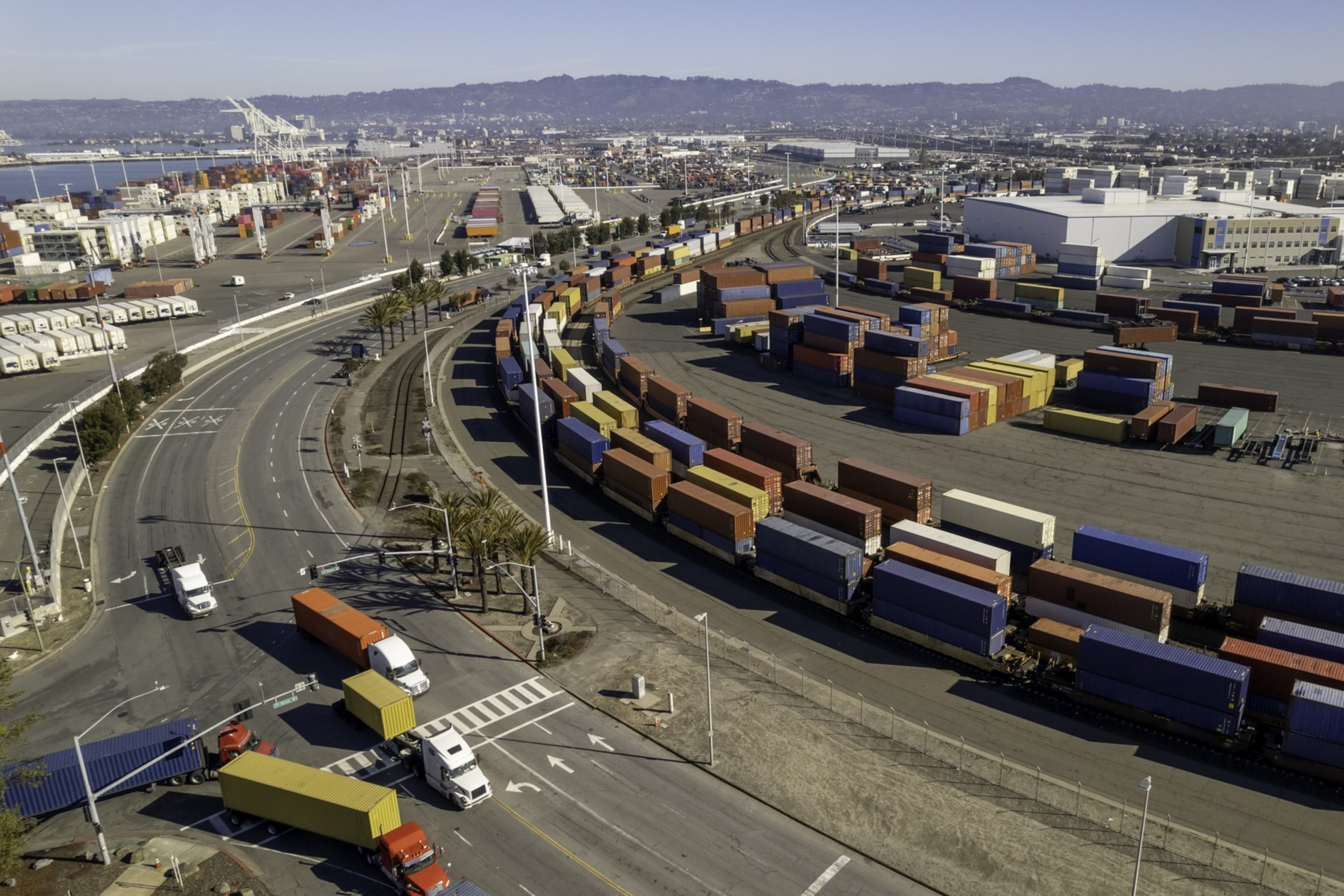 The image shows a large container yard with rows of colorful shipping containers. Trucks navigate the roads around the facility, set against an urban backdrop.