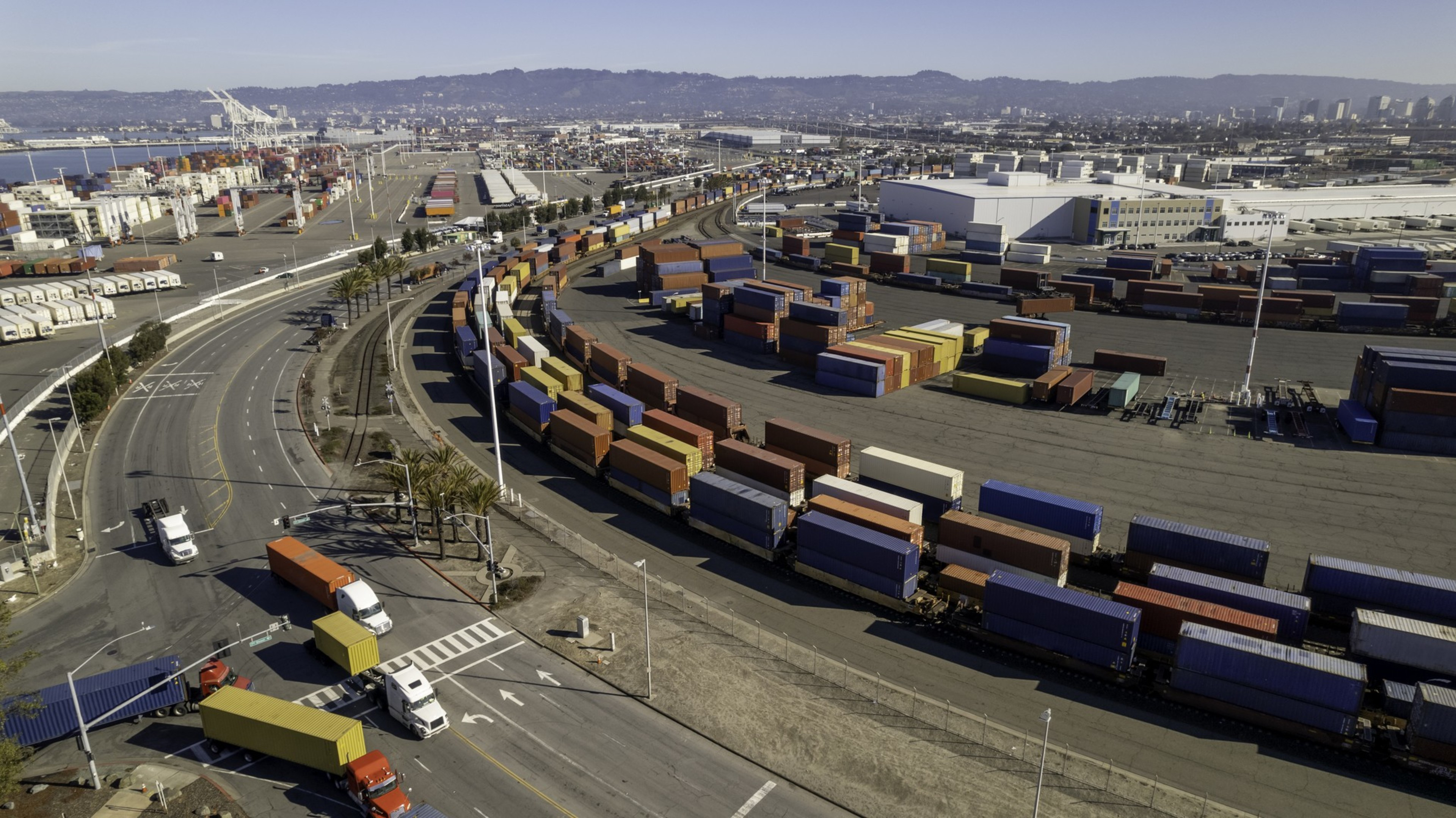 The image shows a large container yard with rows of colorful shipping containers. Trucks navigate the roads around the facility, set against an urban backdrop.