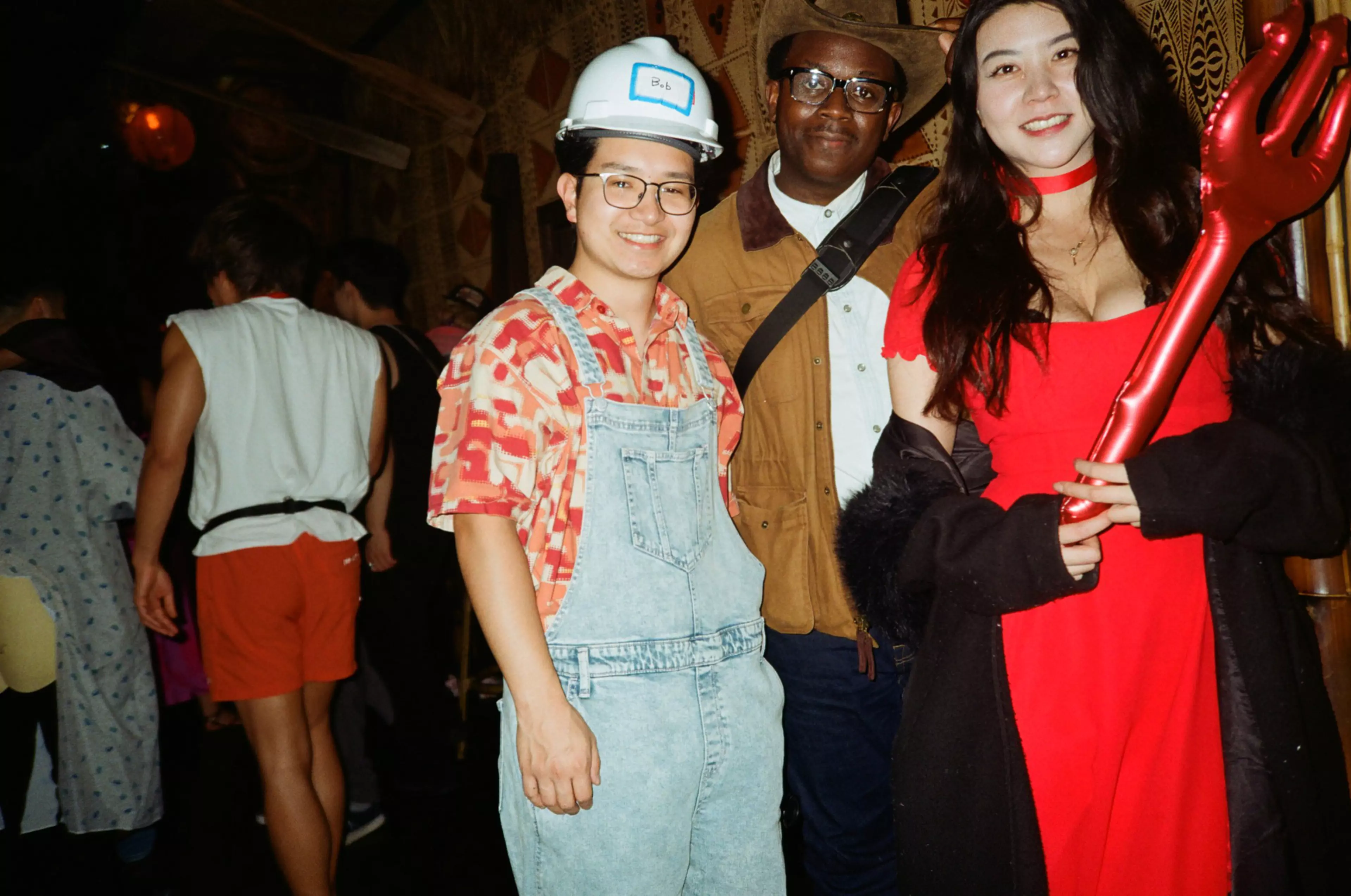 A person in light blue overalls and a hard hat, another in a brown jacket and glasses, and a woman in a red dress holding a red pitchfork pose together.