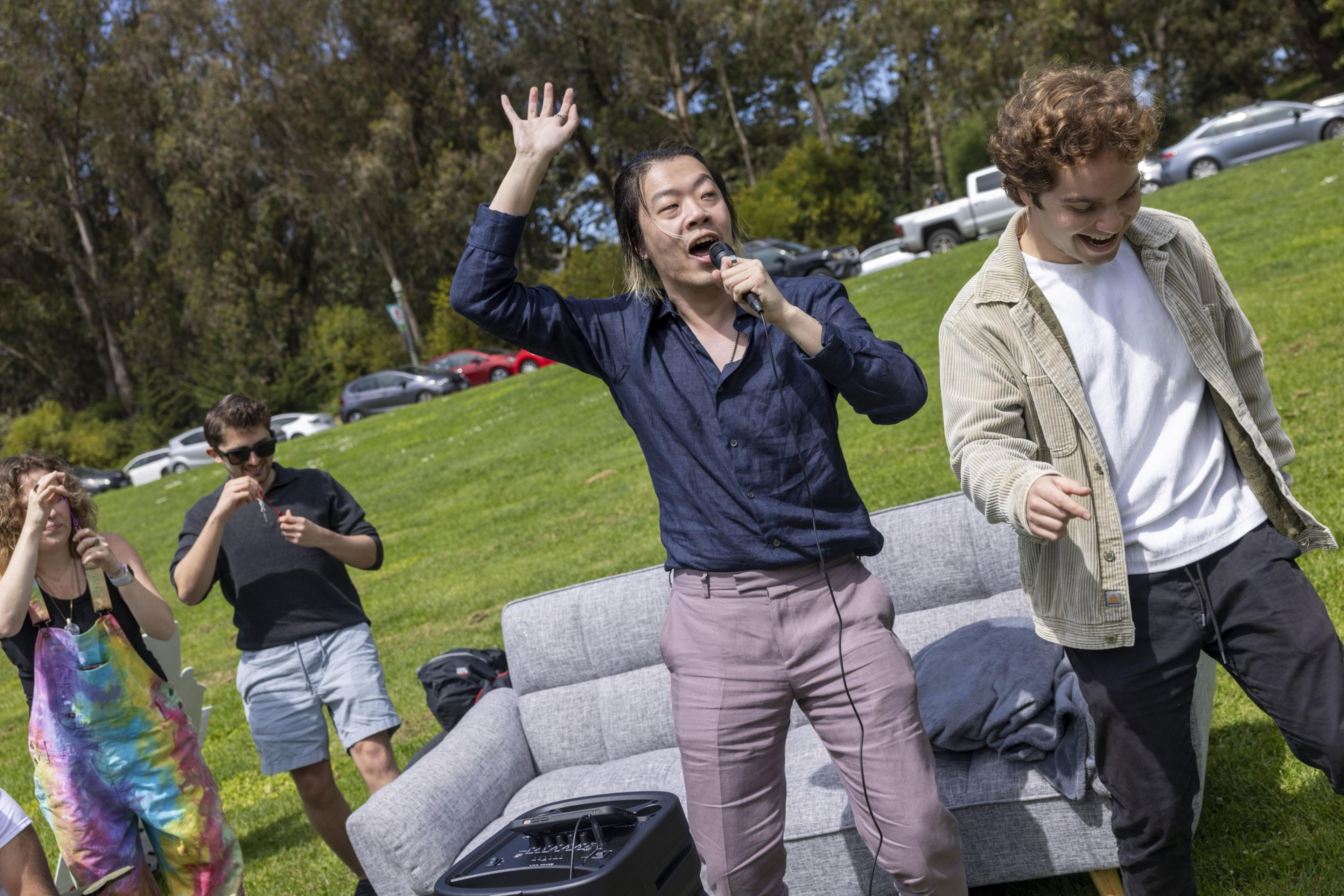 Two people are joyfully singing with a microphone outdoors, while others stand nearby on a grassy field with trees and parked cars in the background.