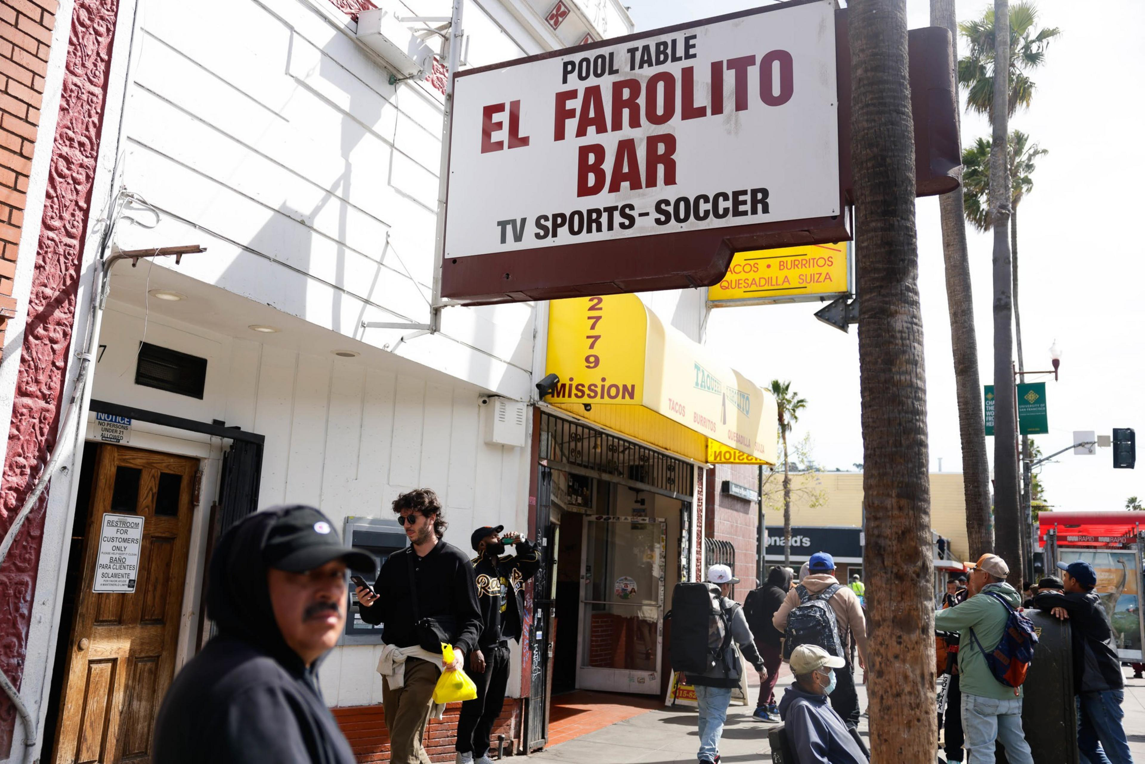 A bustling street corner with a bar sign and pedestrians, palm trees in the background.