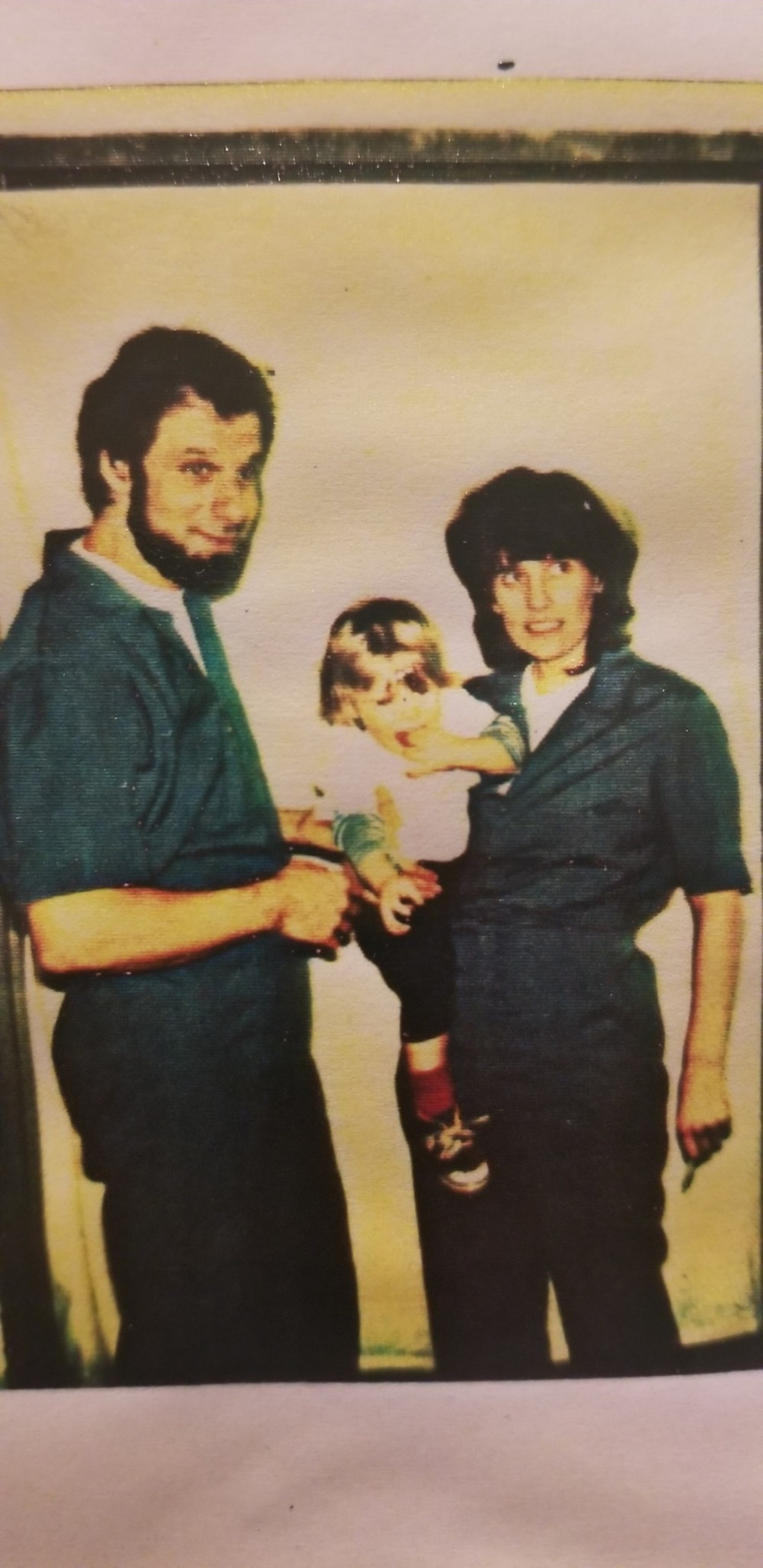 A man and a woman, both in matching outfits, stand side by side holding a small child in their arms, against a light-colored background.