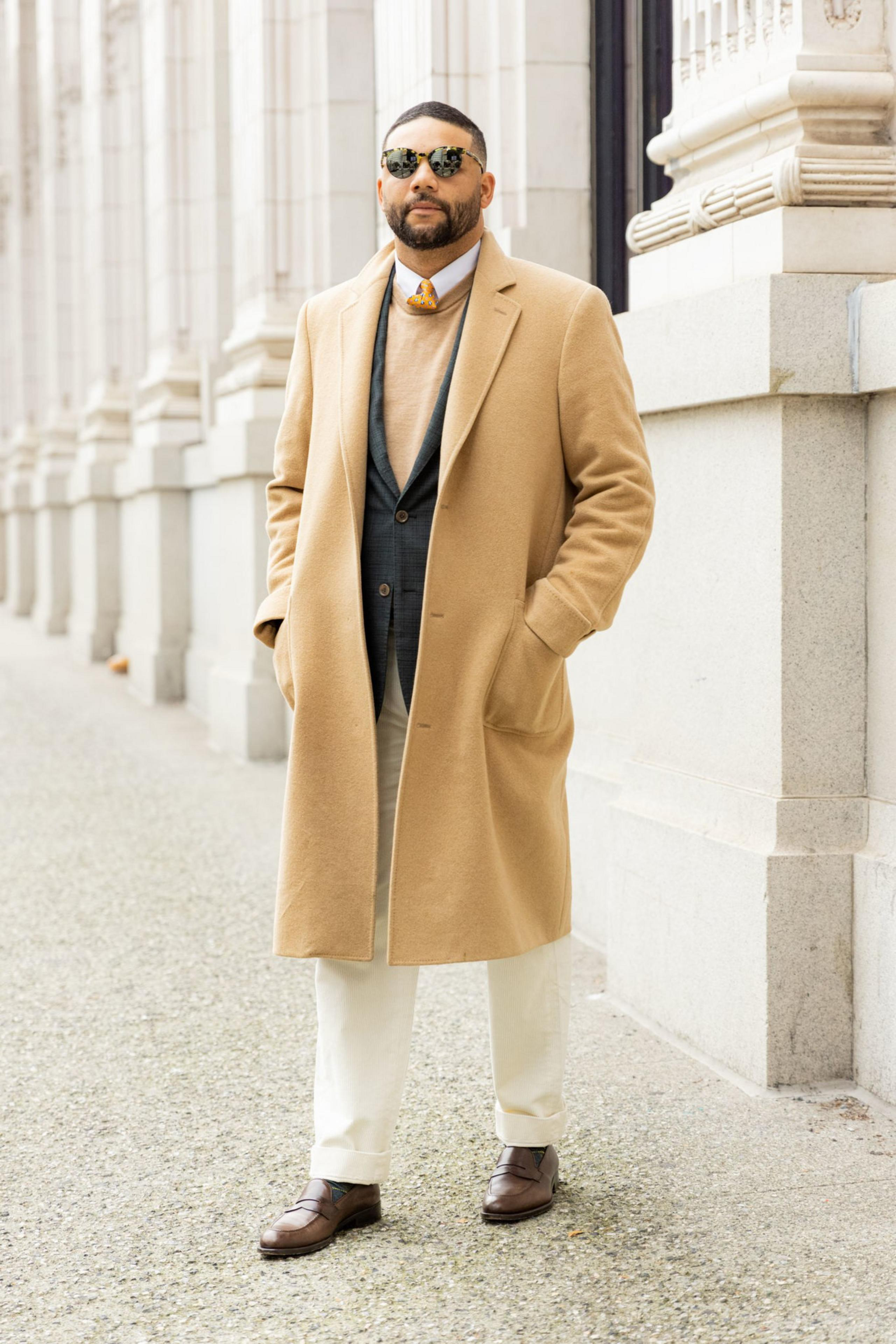 A man stands confidently wearing sunglasses, a camel overcoat, a navy blazer, a tan sweater, white pants, and brown dress shoes, beside a stone columned building.