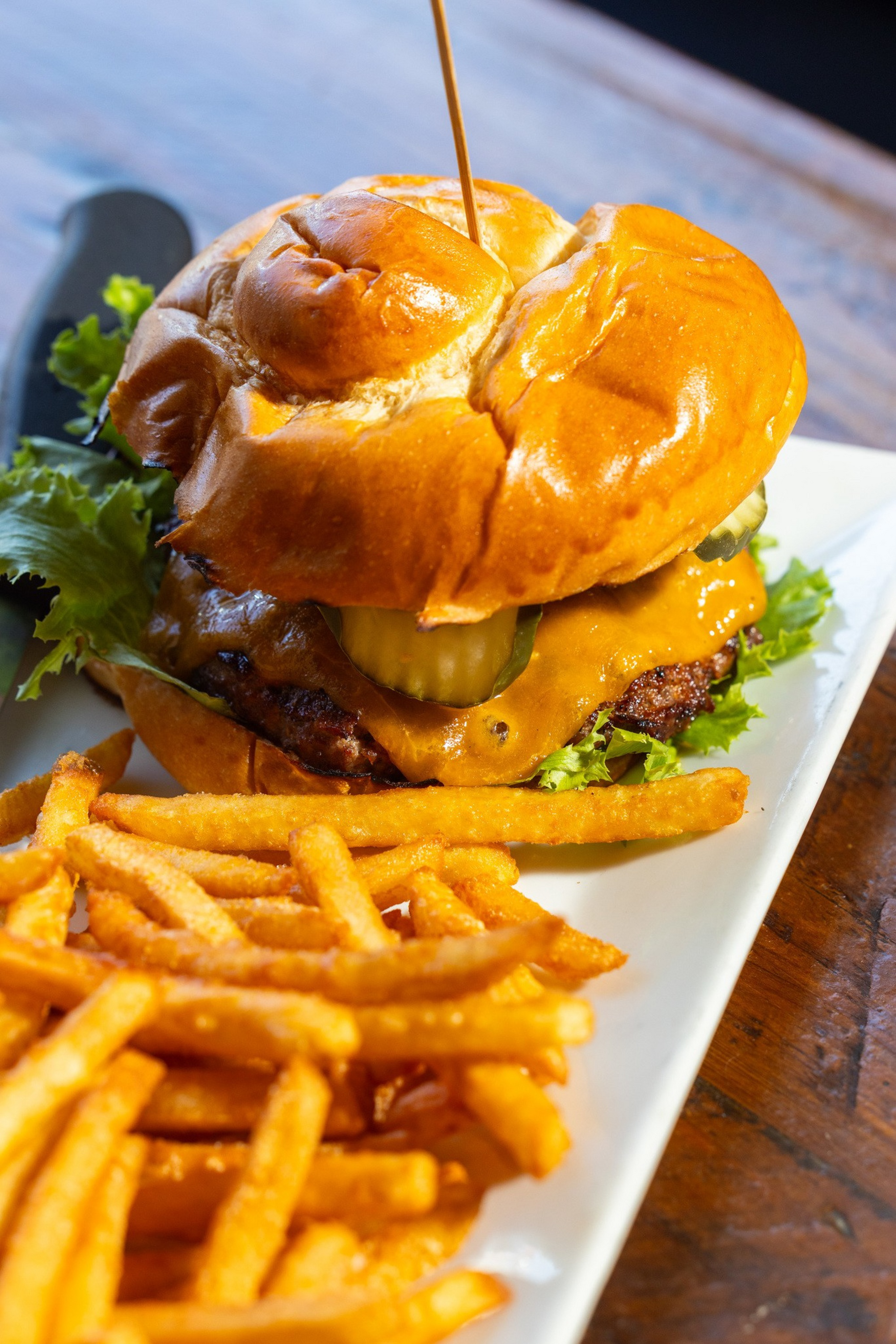 A cheeseburger with lettuce and pickles on a brioche bun is served on a white plate with a large portion of French fries.