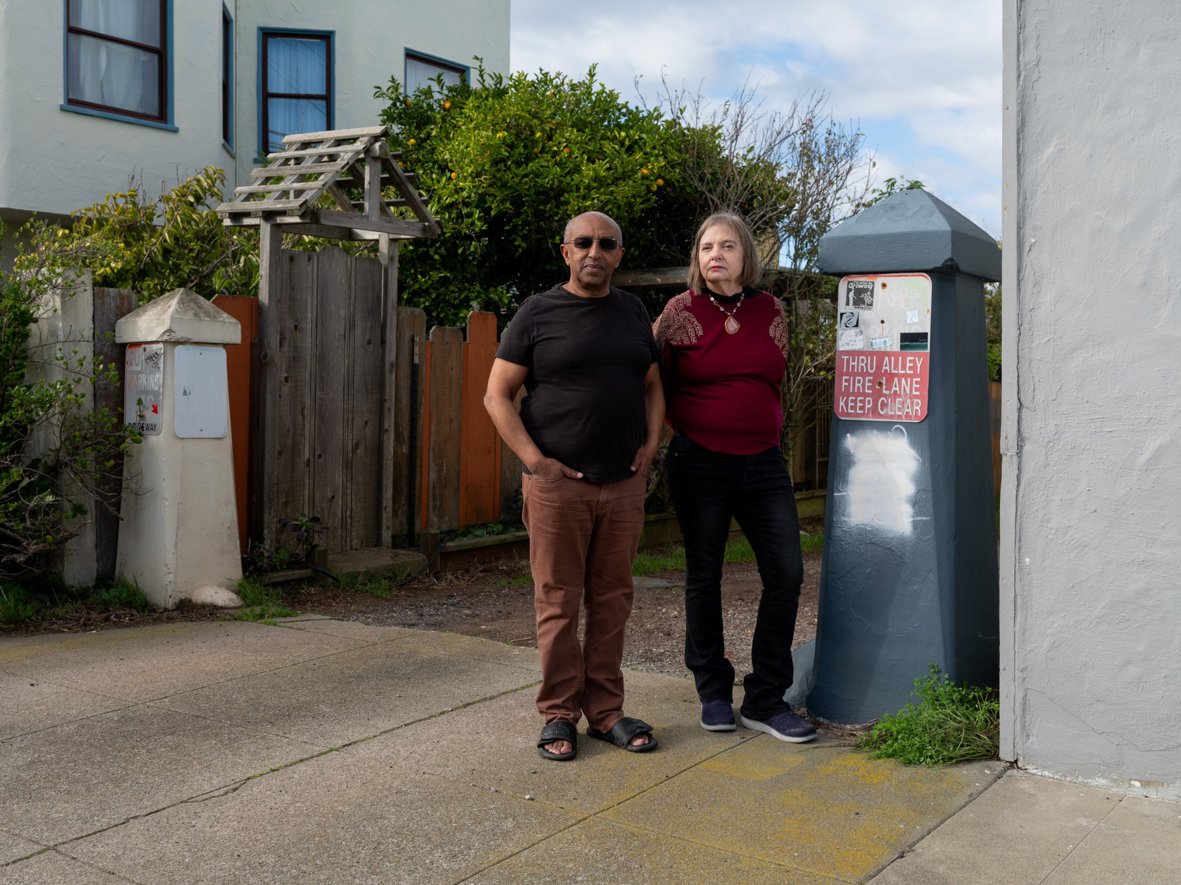 A man in sunglasses and a black shirt stands next to a woman in a burgundy top by a gray post with a red "Fire Lane" sign near a wooden fence.