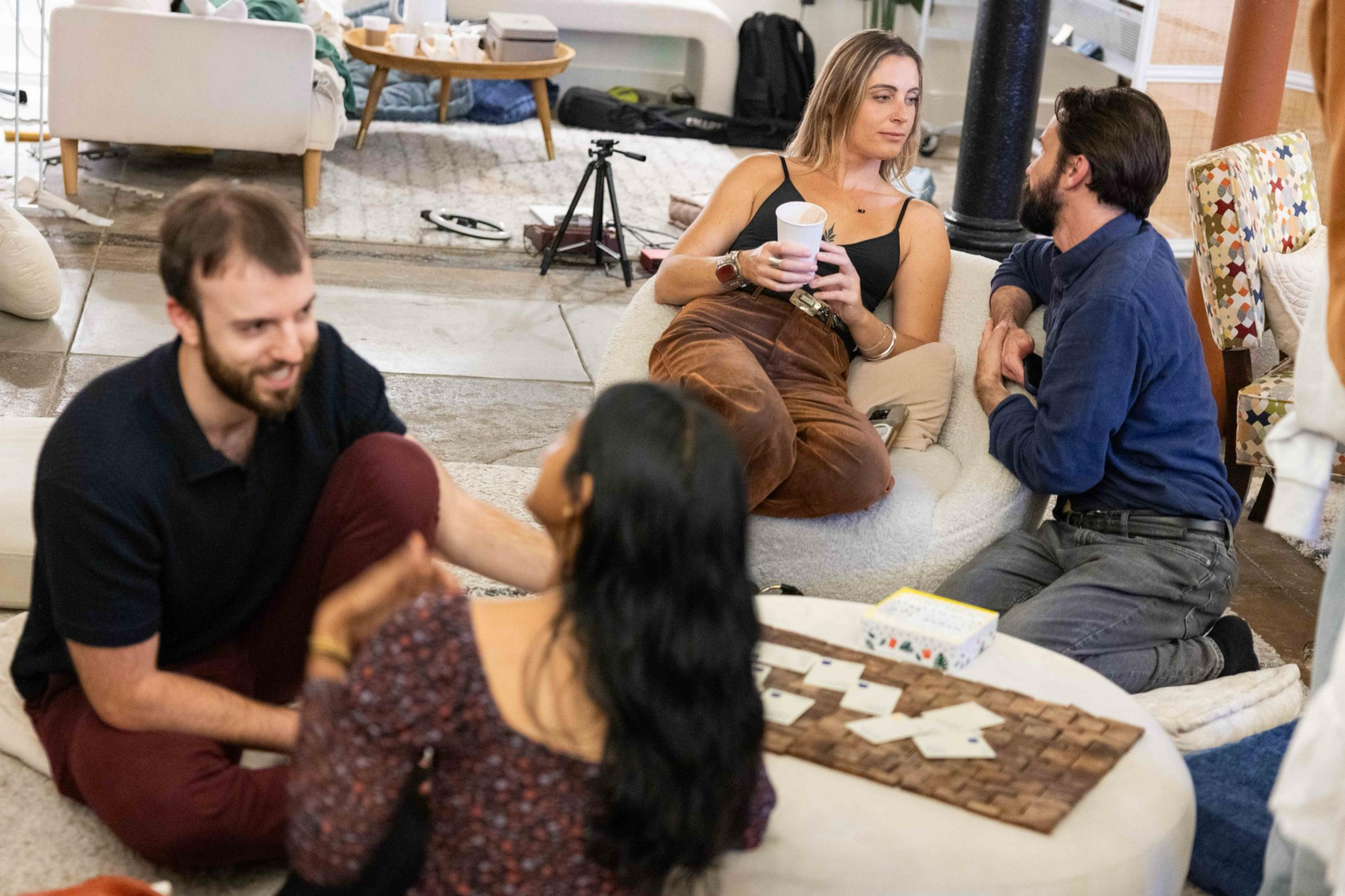 Four people sit on cushions around a low table, engaging in conversation and holding cups in a cozy, casual indoor setting.